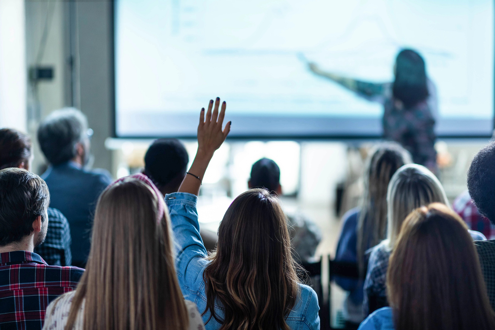 Rear view of a female freelancer raising her hand to ask a question on a seminar in board room.