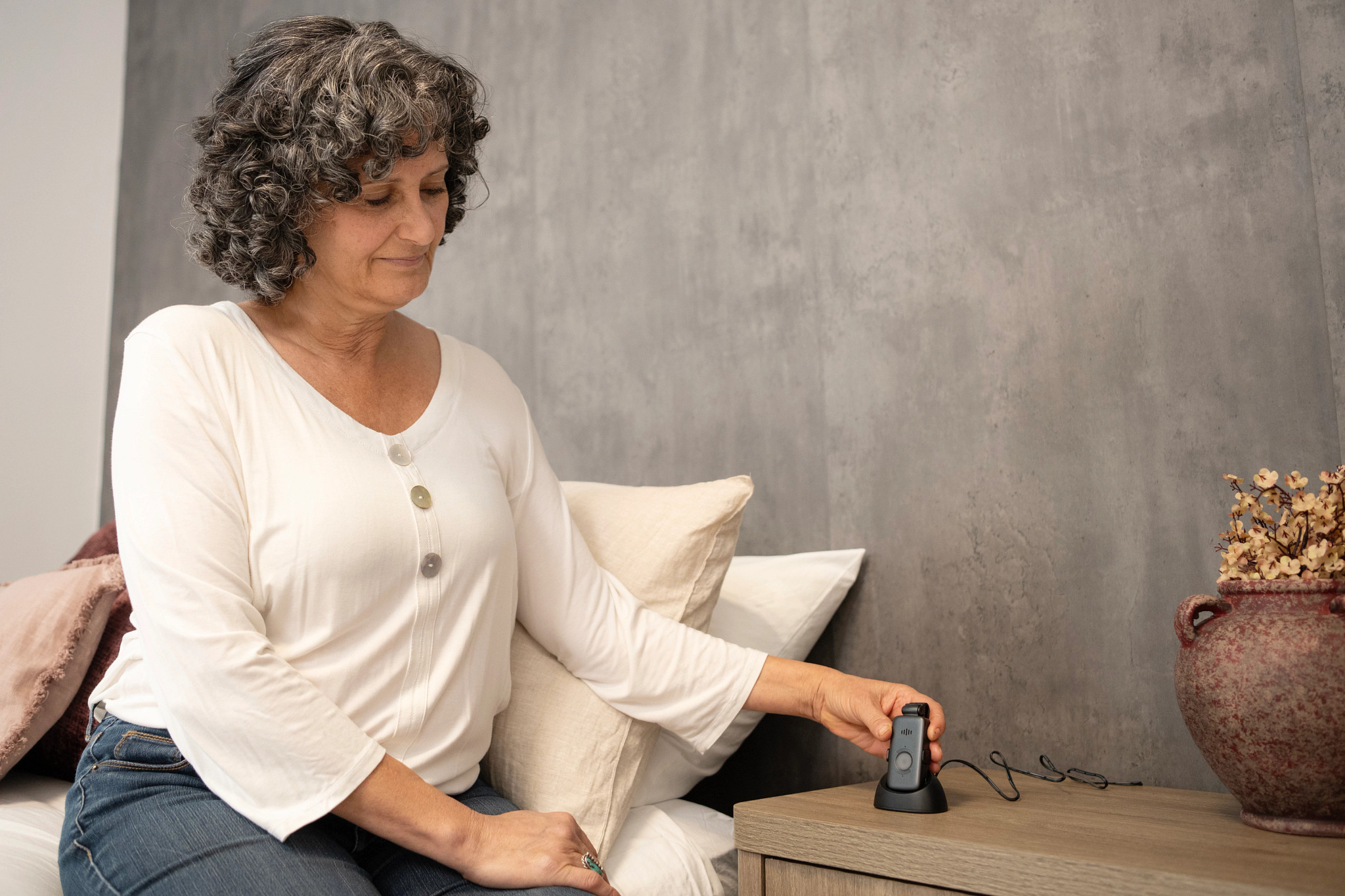 A woman sitting on the edge of a bed with her hand on a device on top of a bedside table
