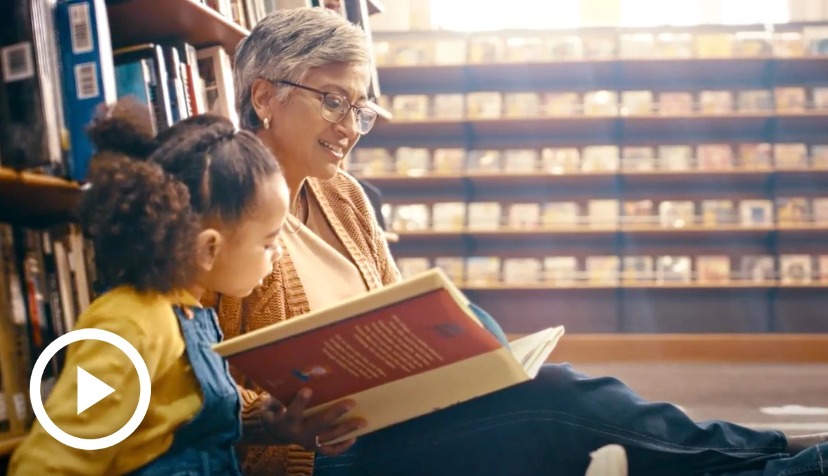 Photograph of an older woman sitting on the floor of a library tutoring a child to read