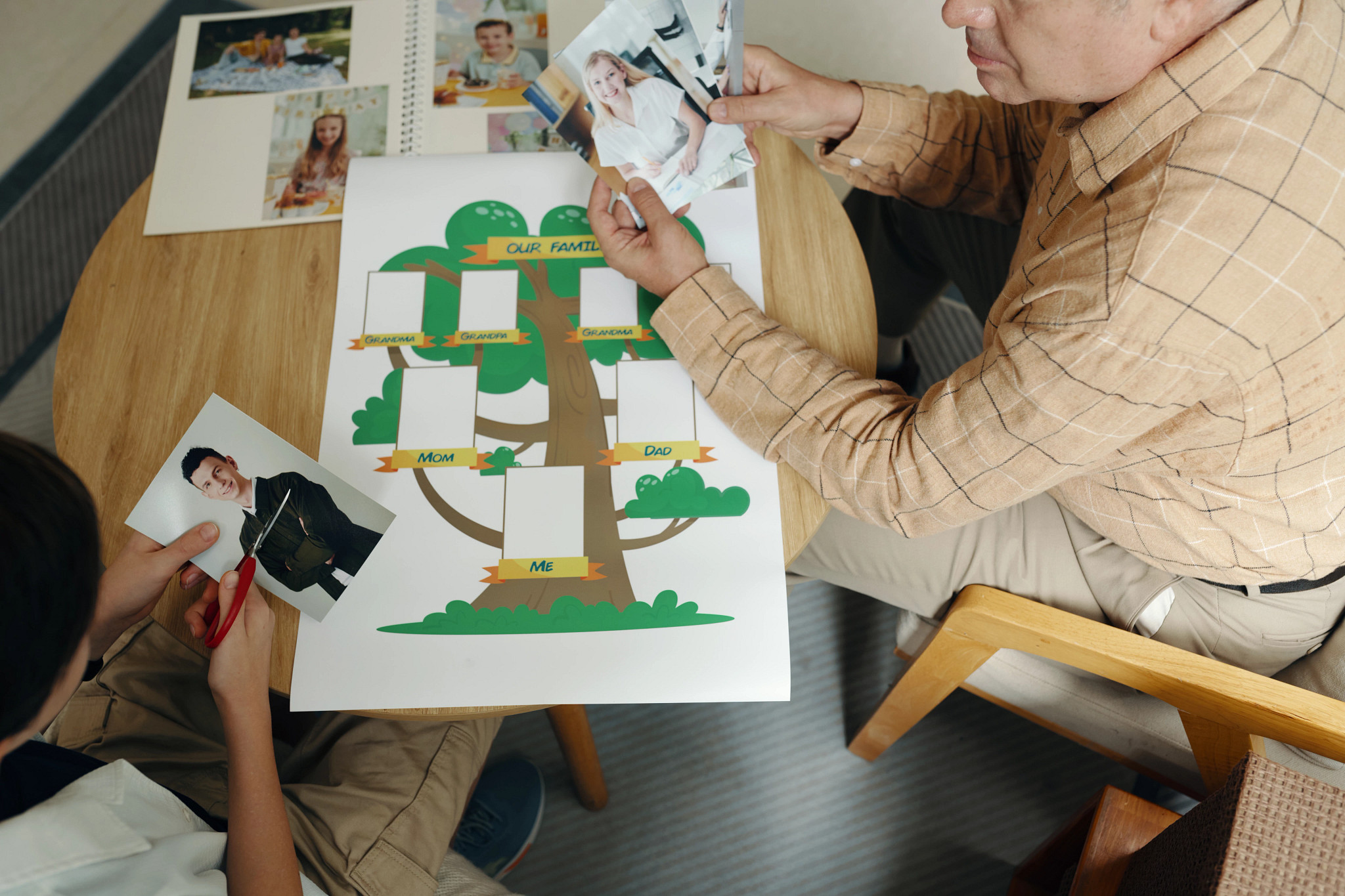 a photo shows an older adult male and a child are seated at a wooden, cutting photos and adding them to a family tree