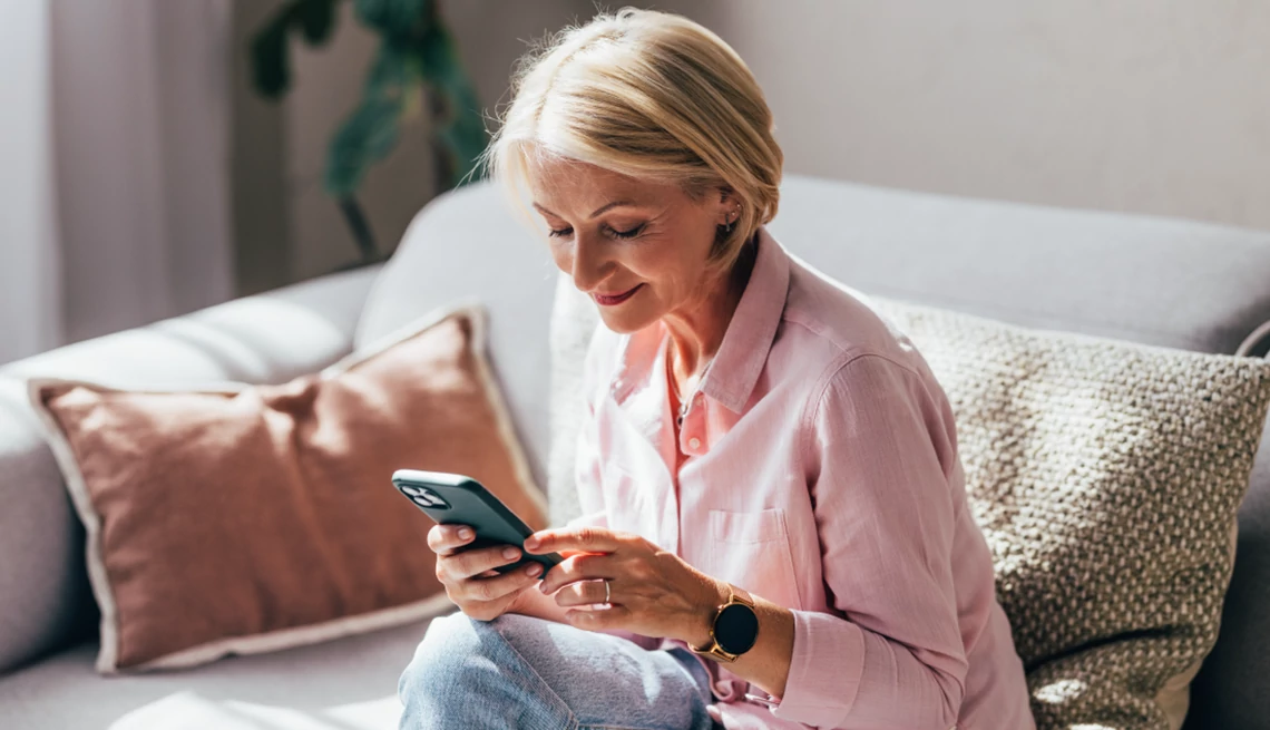 Consumer Cellular Woman sitting on a sofa looking at her smartphone and smiling in the morning light