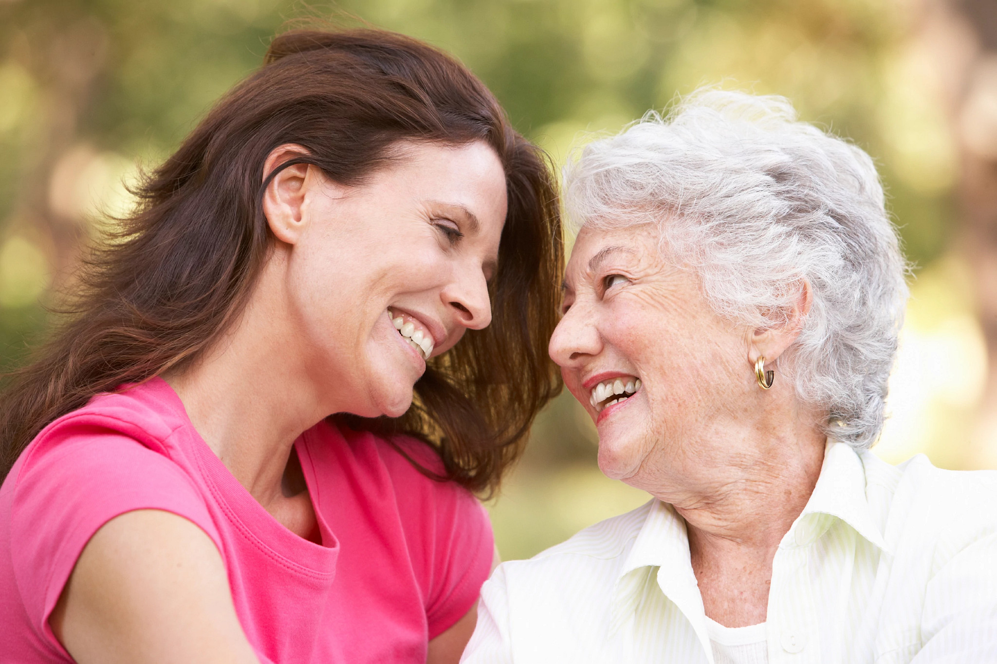 A close-up of a younger woman with brown hair and an older woman with short gray hair smiling and looking at each other outdoors.