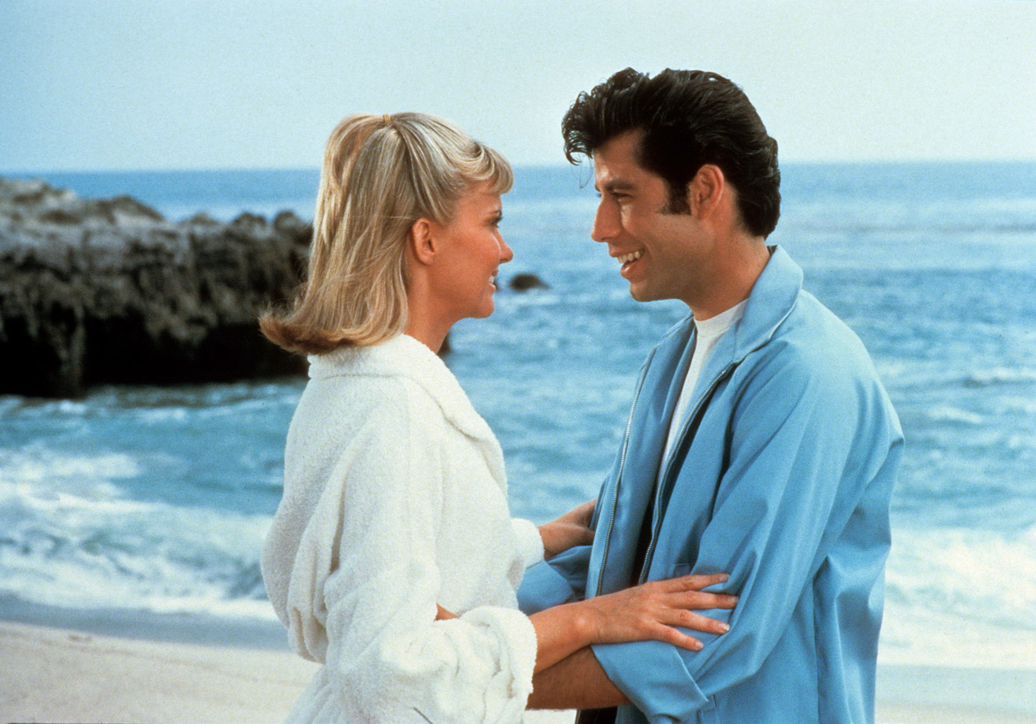 Olivia Newton-John and John Travolta standing close together on a beach with ocean waves and rocky shoreline in the background in the film Grease