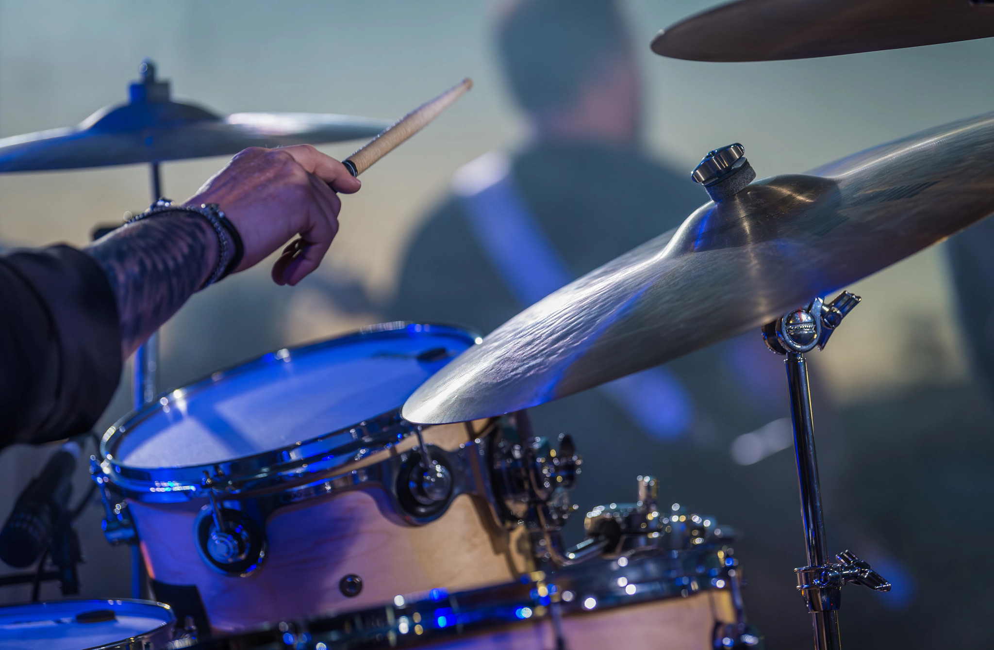 Blue lights glow on a drum set as a drummer prepares to hit a cymbal with his drumstick during a live concert