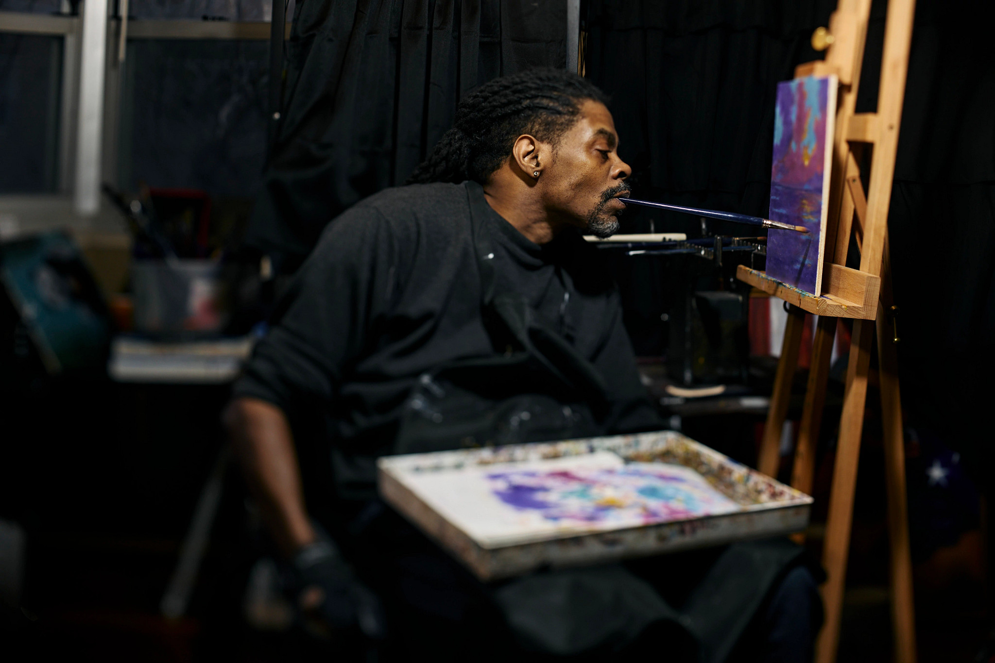antonio davis using his mouth to hold a paintbrush while working on a painting on an easel