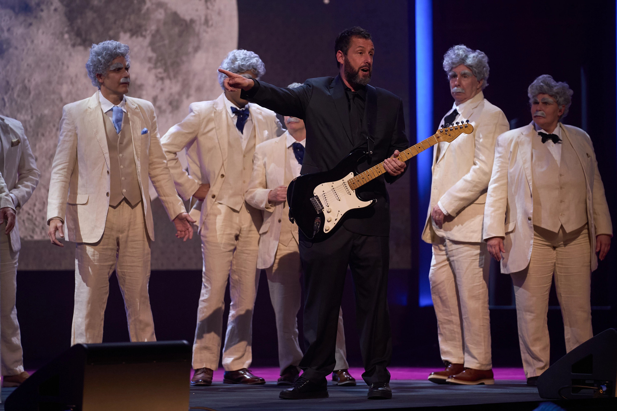 adam sandler holds a guitar in front of a group of people dressed as mark twain
