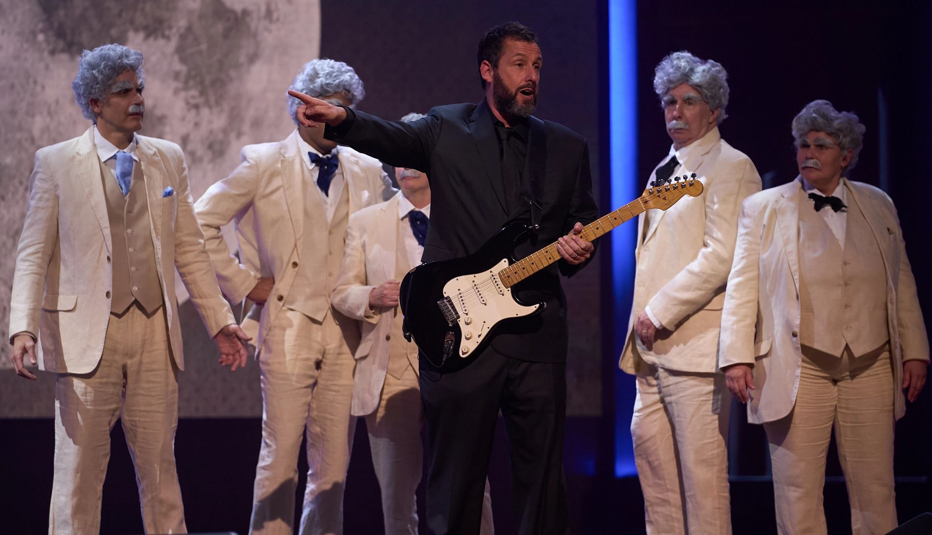 Adam Sandler adam sandler holds a guitar in front of a group of people dressed as mark twain