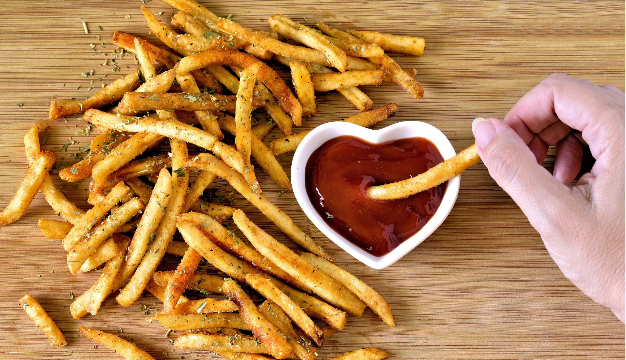 National French Fry Day a photo with an overhead viewpoint shows a woman partaking from a cutting board filled with French fries seasoned with salt and herbs. She is a dipping a fry into a heart-shaped ramekin filled with ketchup.