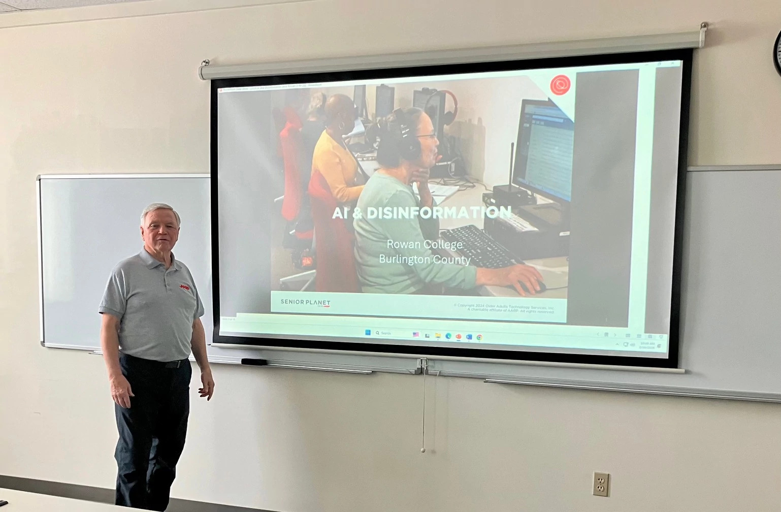 An instructor standing next to a large projector screen displaying a presentation titled "Ai & Disinformation" for the Rowan College at Burlington County Life Program.