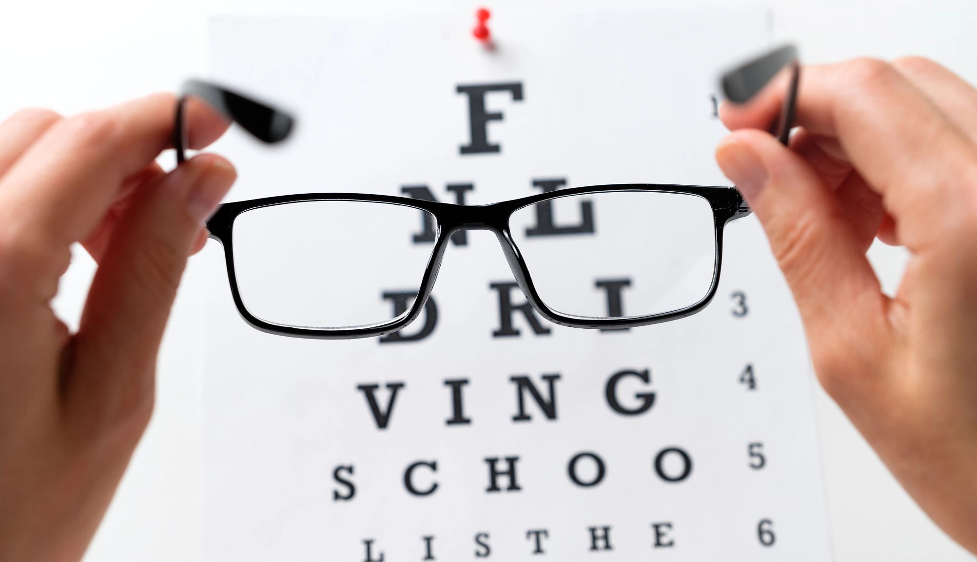 Where to Start Eyeglasses in the hands of someone, in front of the Snellen eye chart