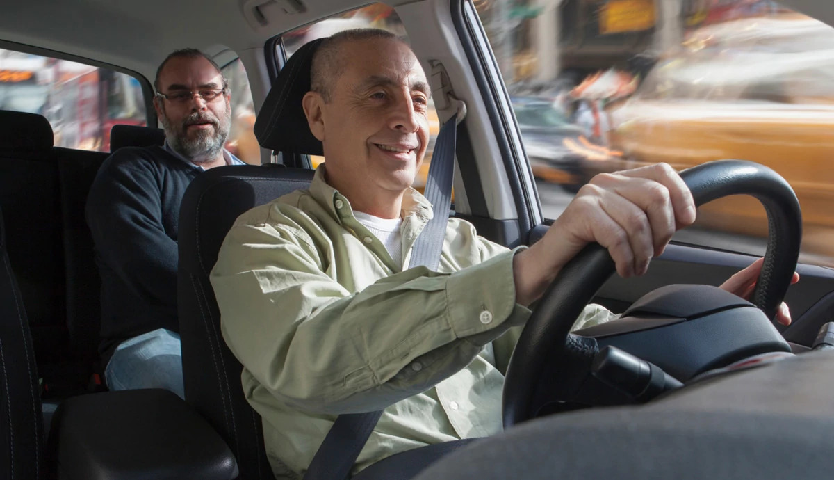 Two men in a car. One is in the driver's seat with both hands on the steering wheel, the other is riding behind him in the back seat.