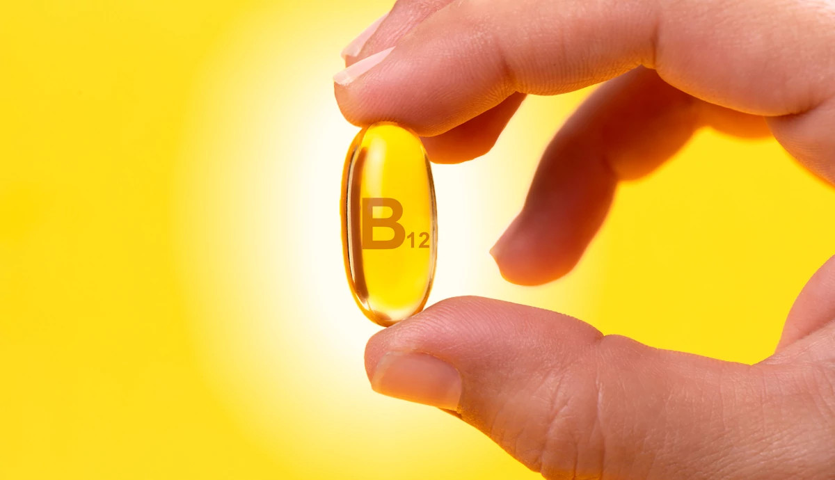 close-up shot on a bright yellow background shows a person's thumb and forefinger holding a single, translucent golden gel capsule labeled "B12."