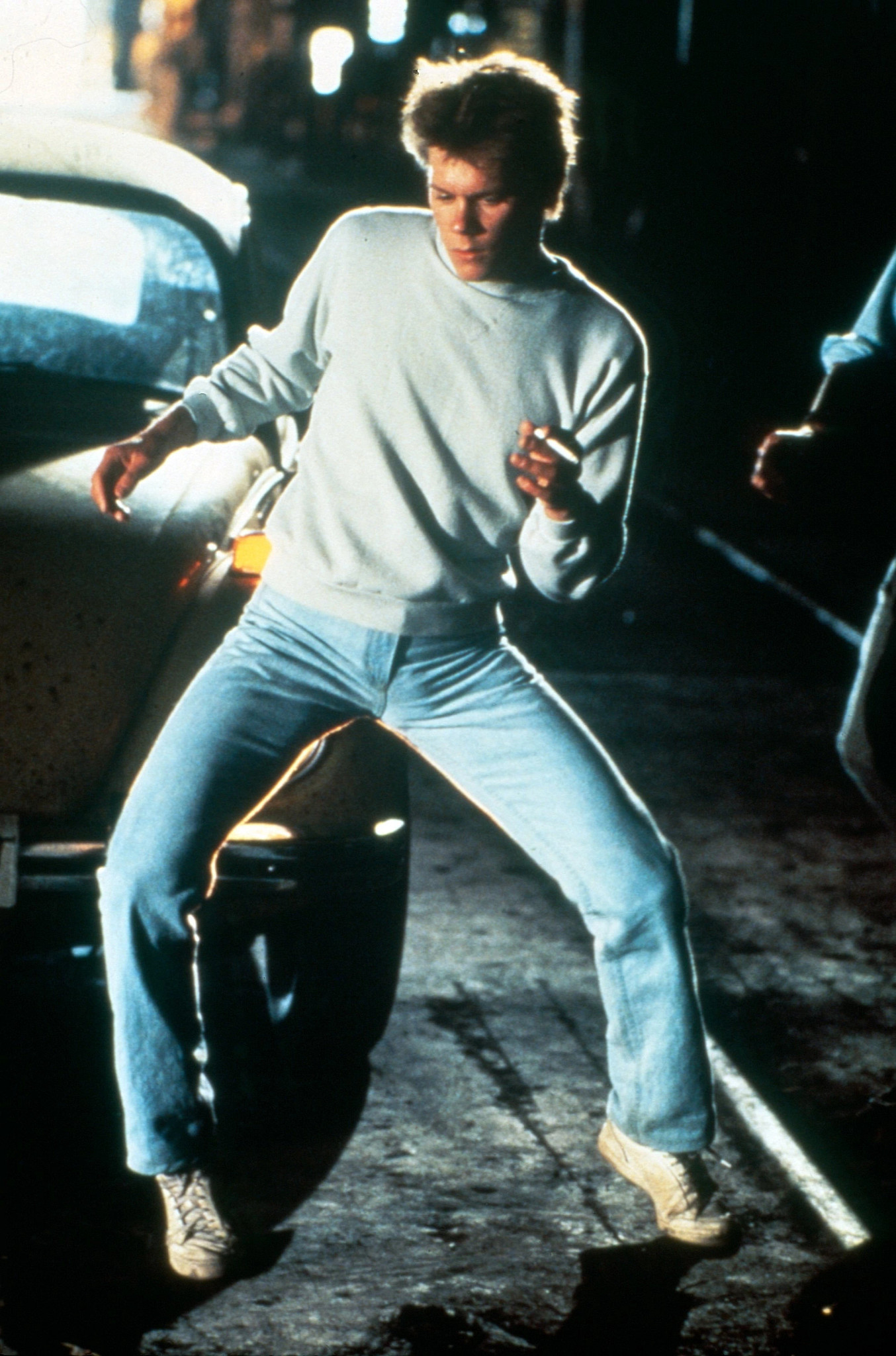 Kevin Bacon dancing energetically on a dimly lit city street beside parked cars at night, wearing casual clothing and illuminated by streetlights in the film Footloose