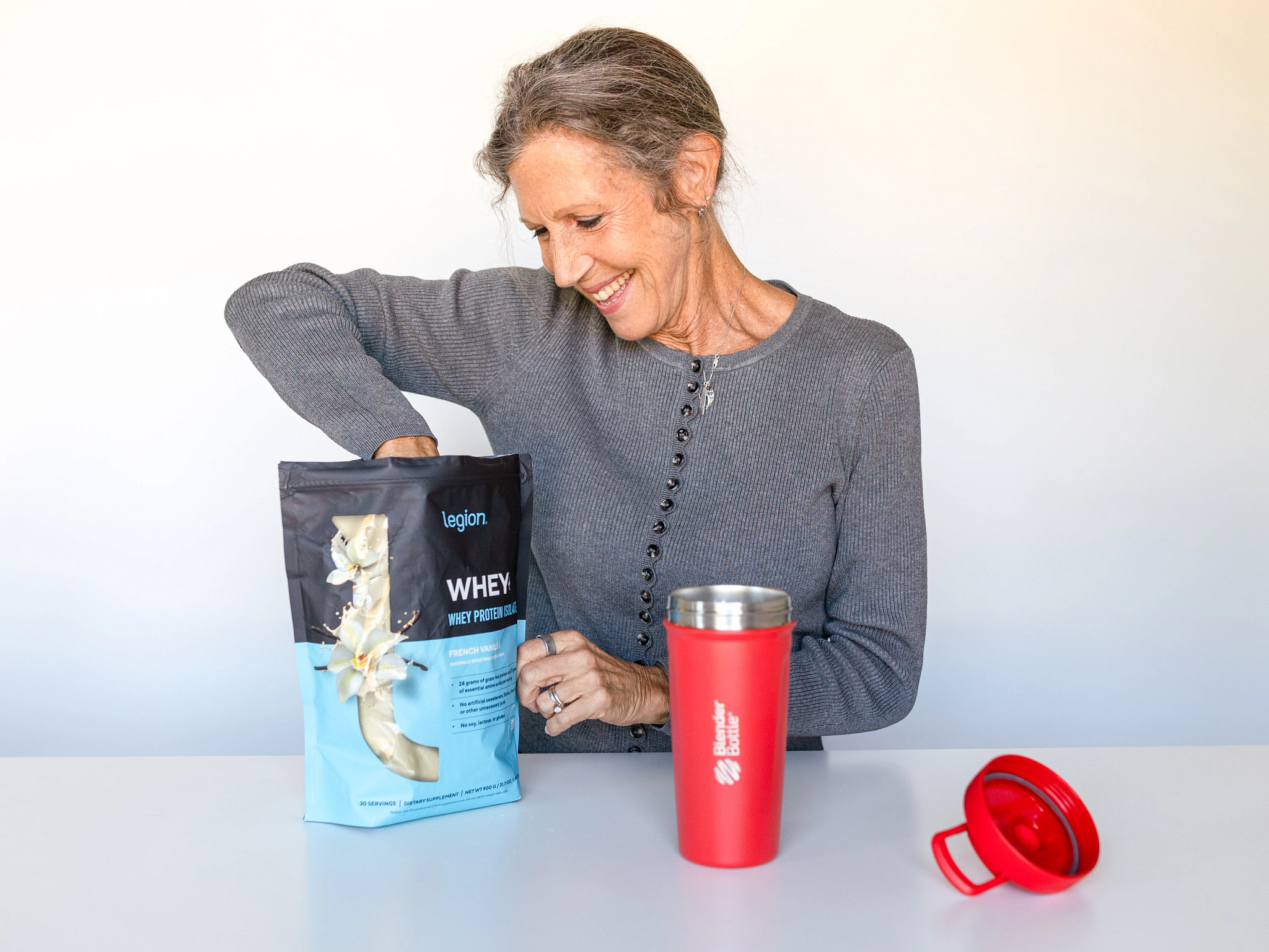 A woman scooping Legion Whey+ Protein Isolate with a red shaker bottle in front 