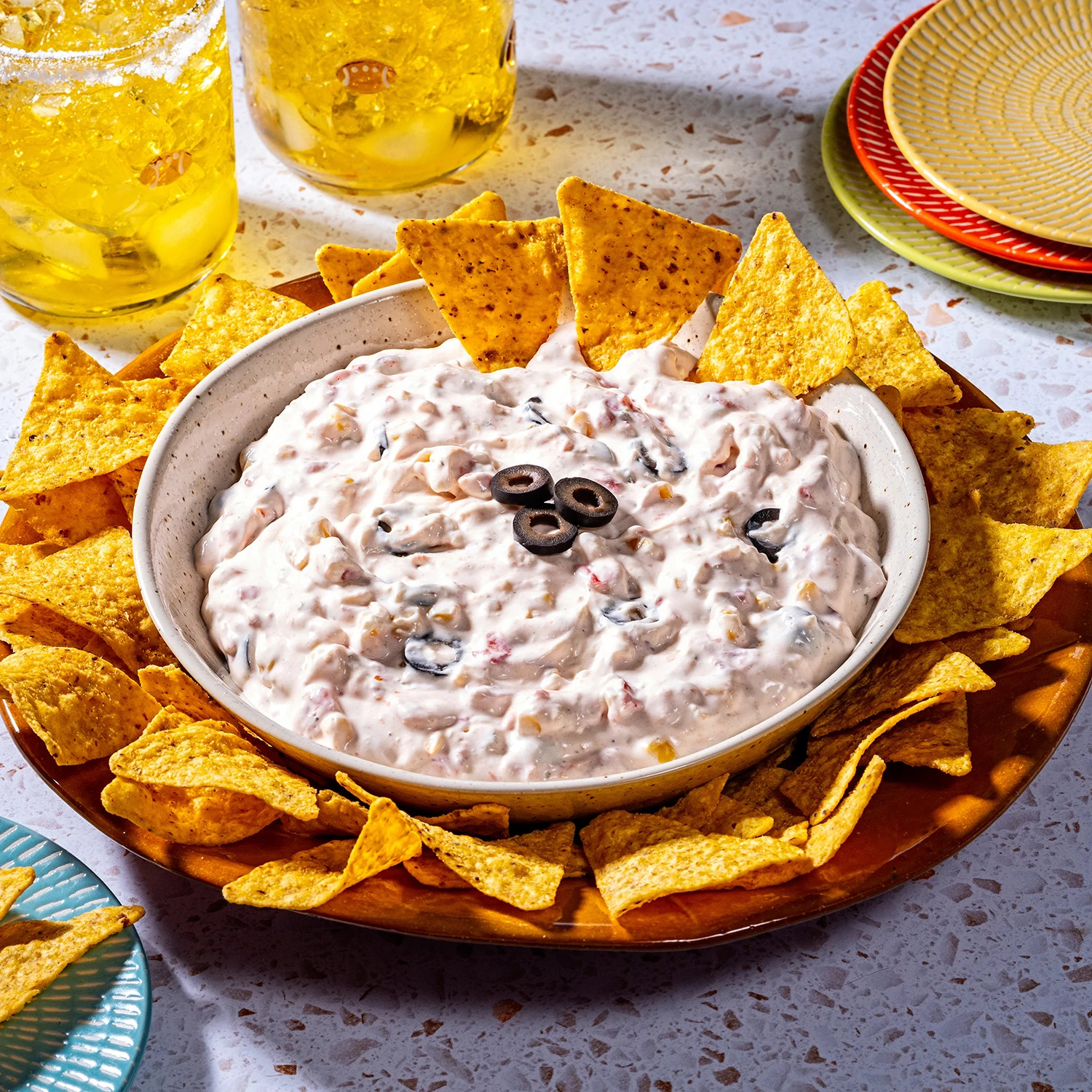 Creamy dip topped with black olives served with tortilla chips on a wooden platter.