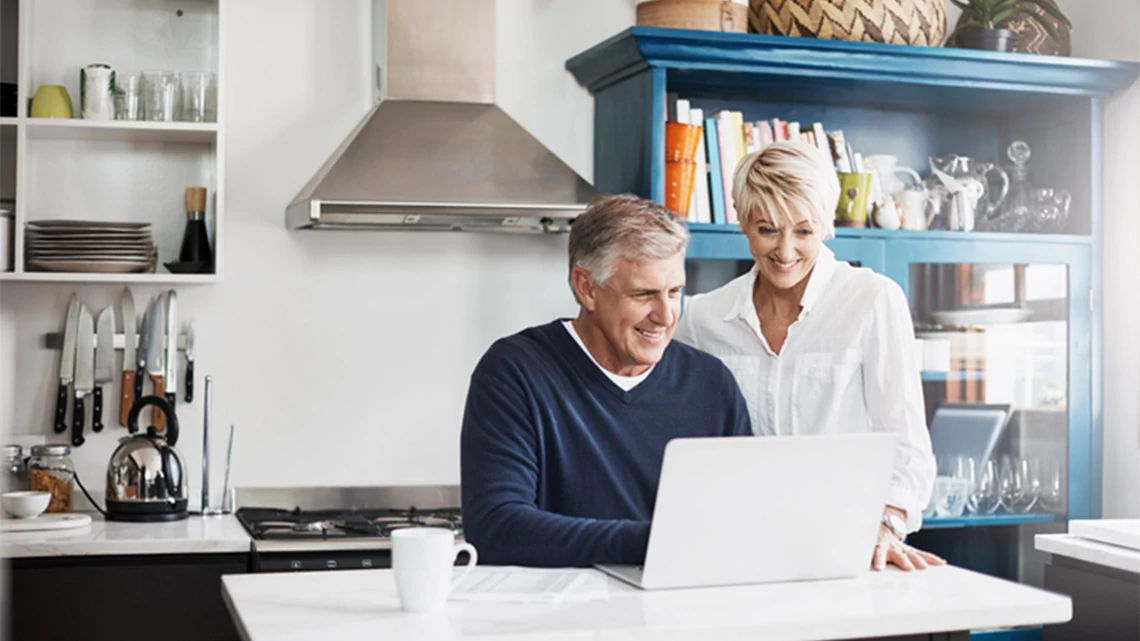 Couple in kitchen, man sitting, woman standing, both looking at a laptop, smiling, coffee cup on island