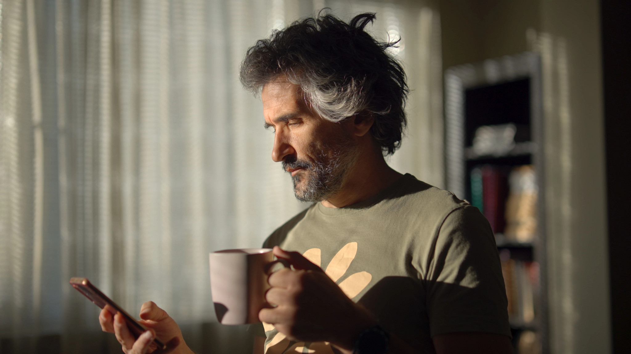 A man holding a coffee mug in one hand and looking at his smartphone in the other inside a home