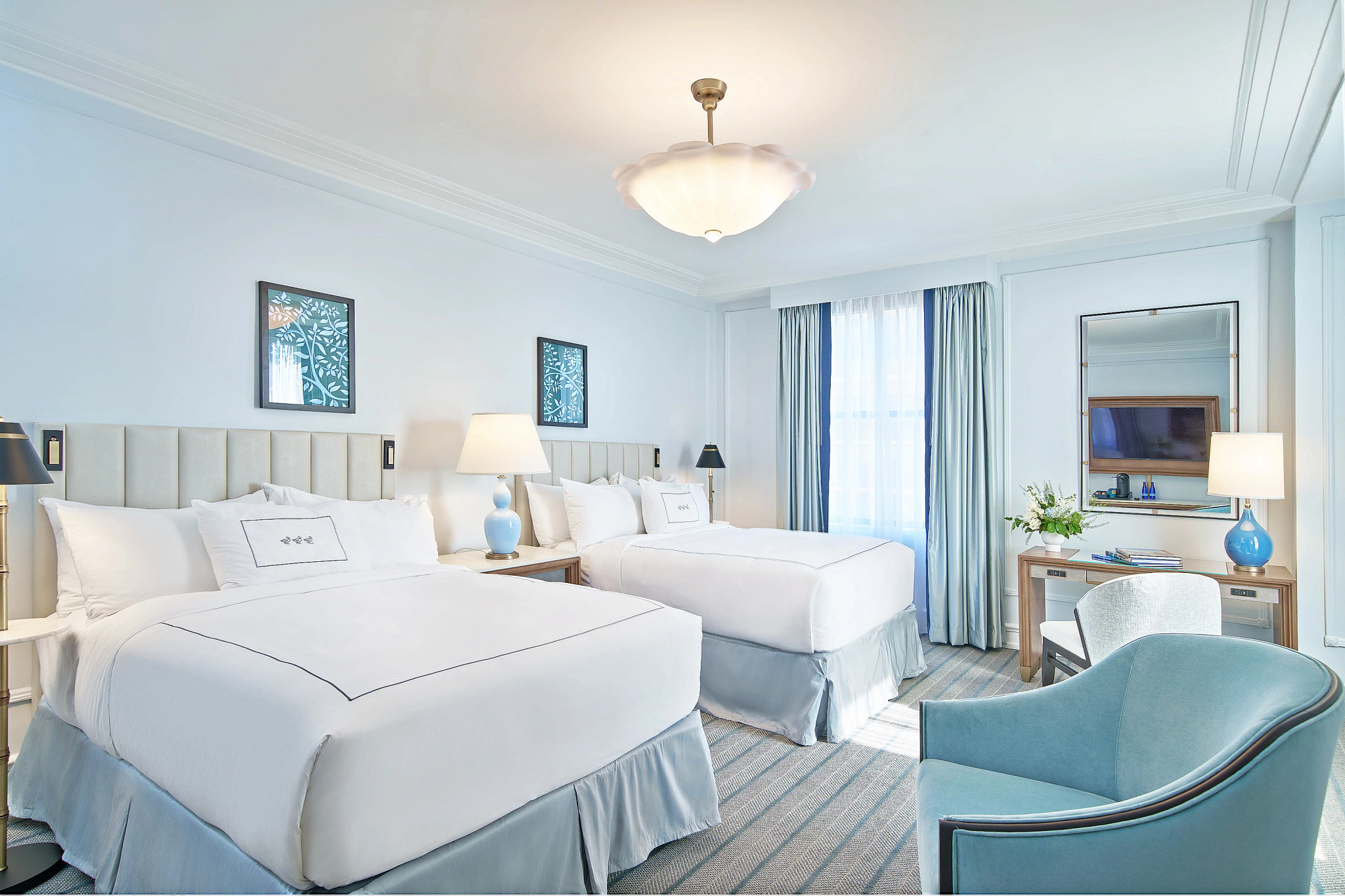 Bright and stylish deluxe hotel guest room featuring two double beds, a seating area and large windows with natural light.