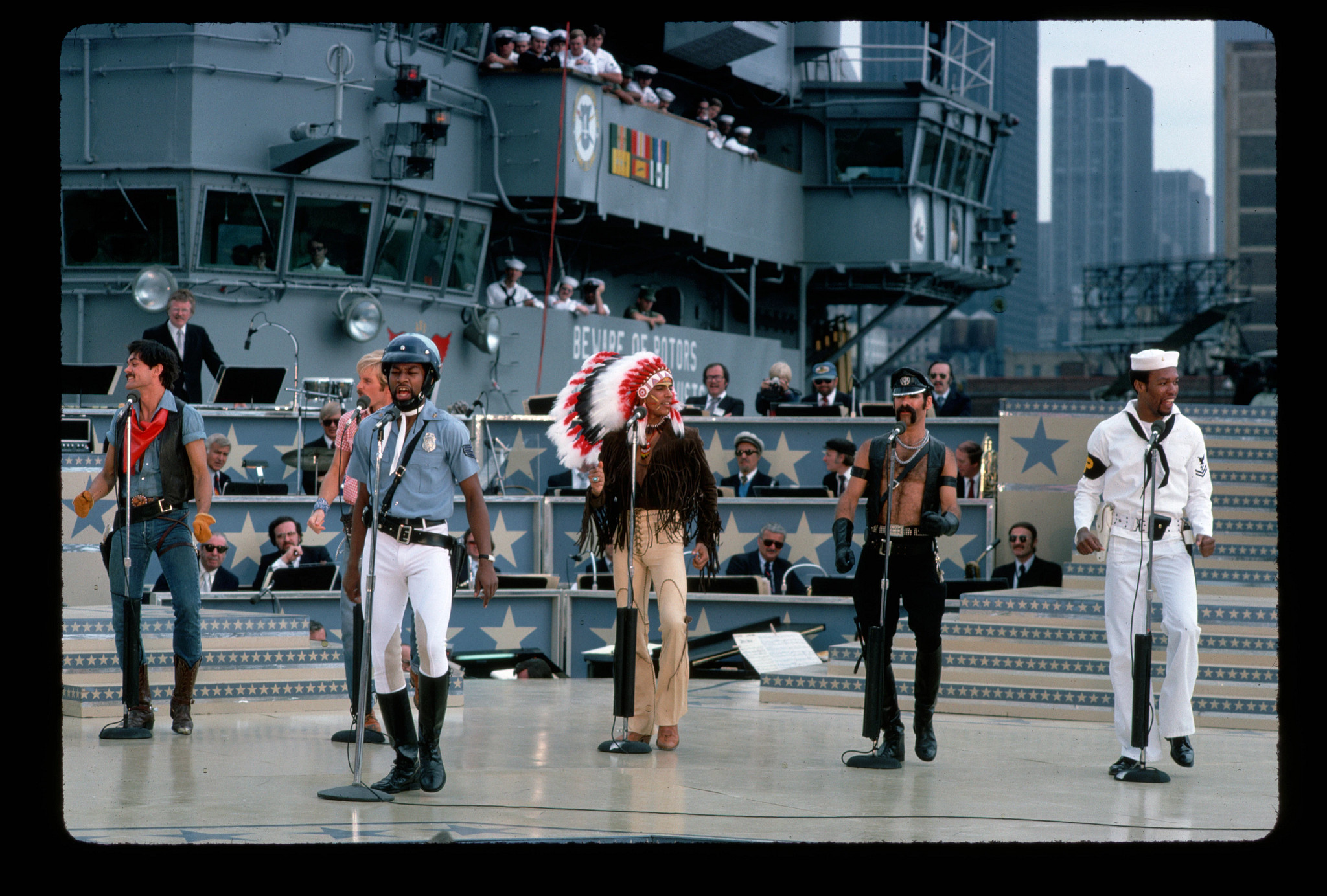 village people singers perform onstage in their costumes as a cowboy, construction worker, police officer, indian chief, biker and sailor