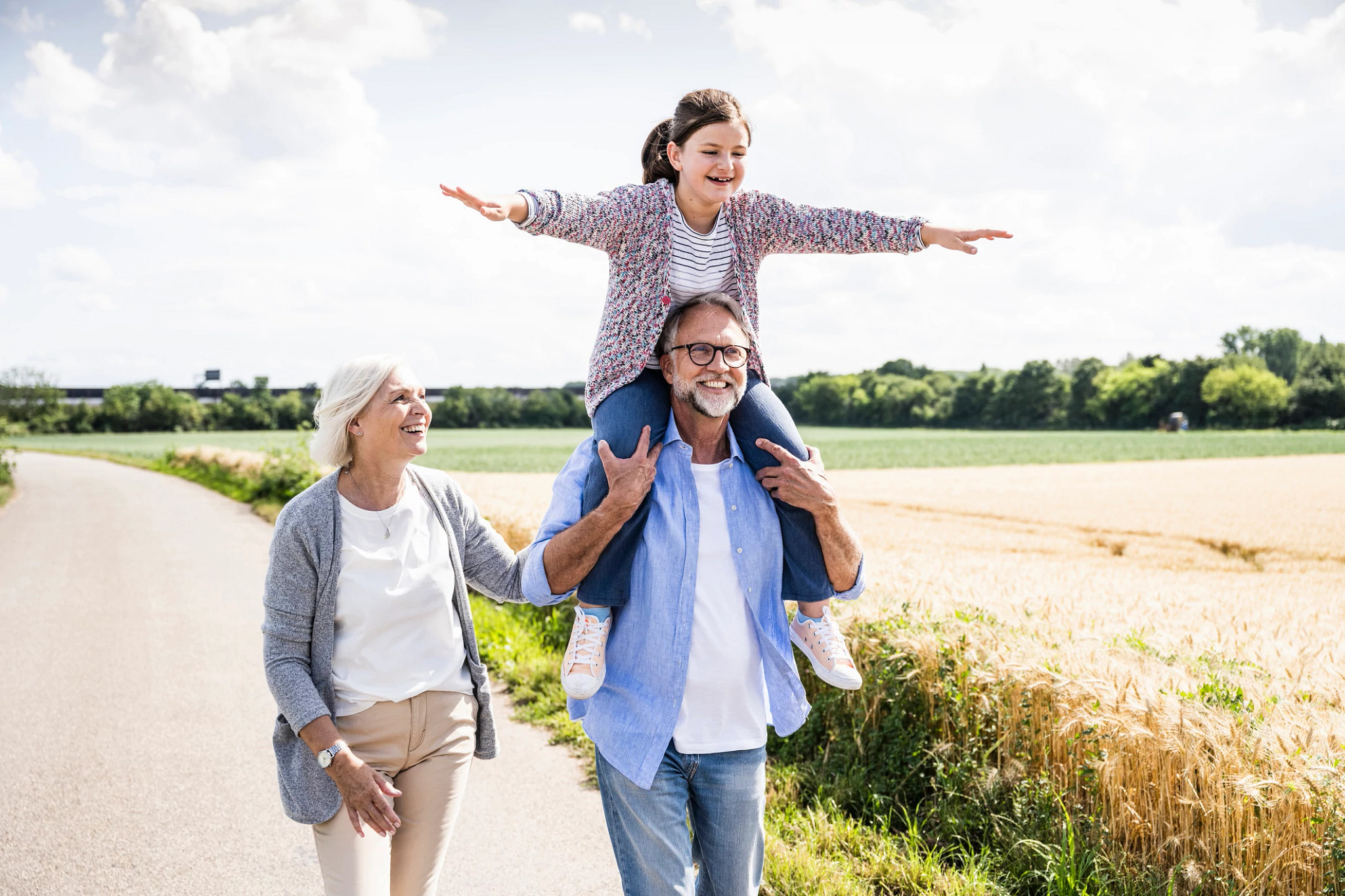 Grandfather carrying cheerful granddaughter on shoulder while walking by woman on road