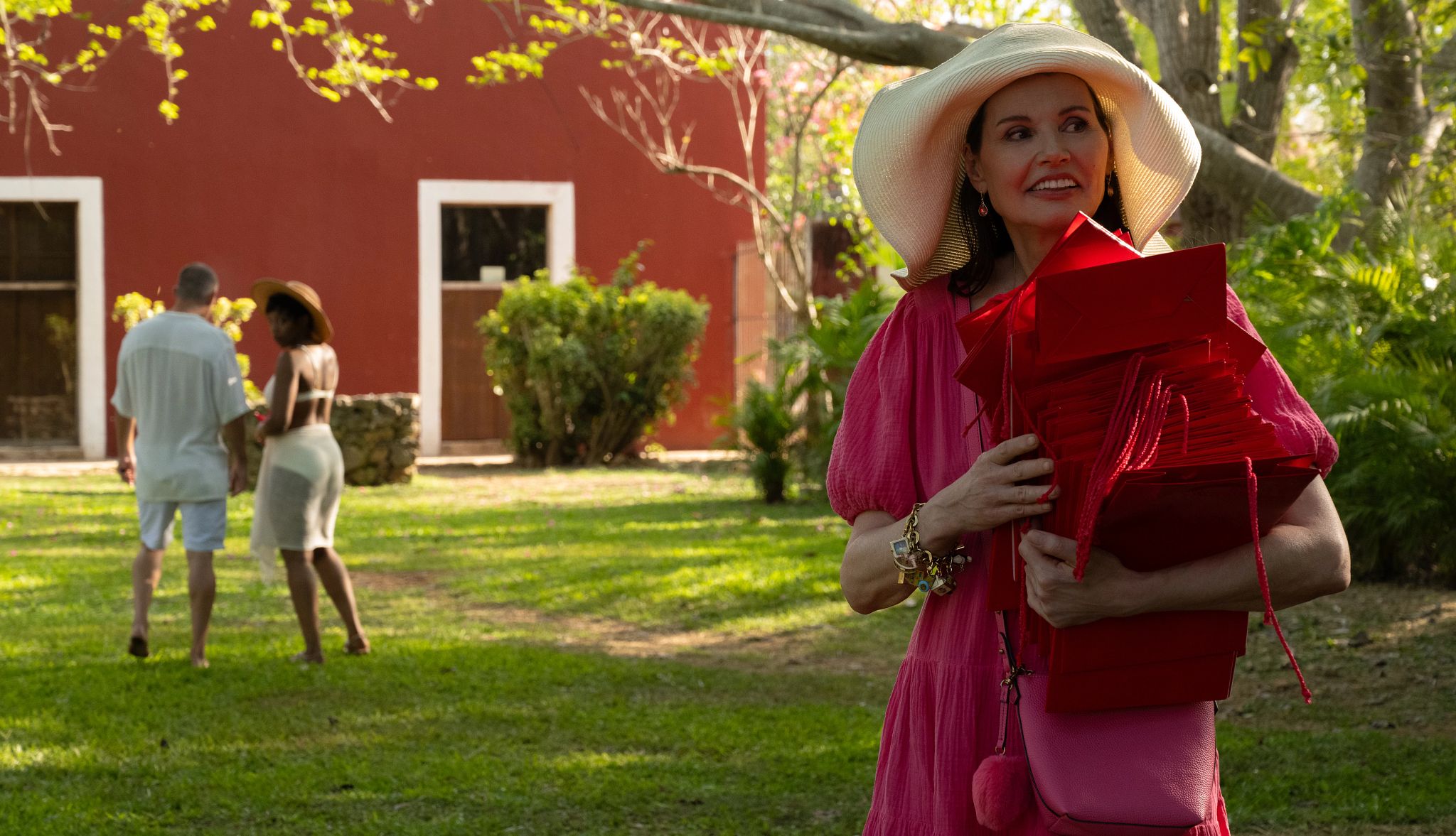 Geena Davis stars with Channing Tatum and Naomi Ackie in 'Blink Twice' Geena Davis standing outside wearing big hat and holding a bunch of red gift bags; Channing Tatum and Naomi Ackie in the background with backs towards her
