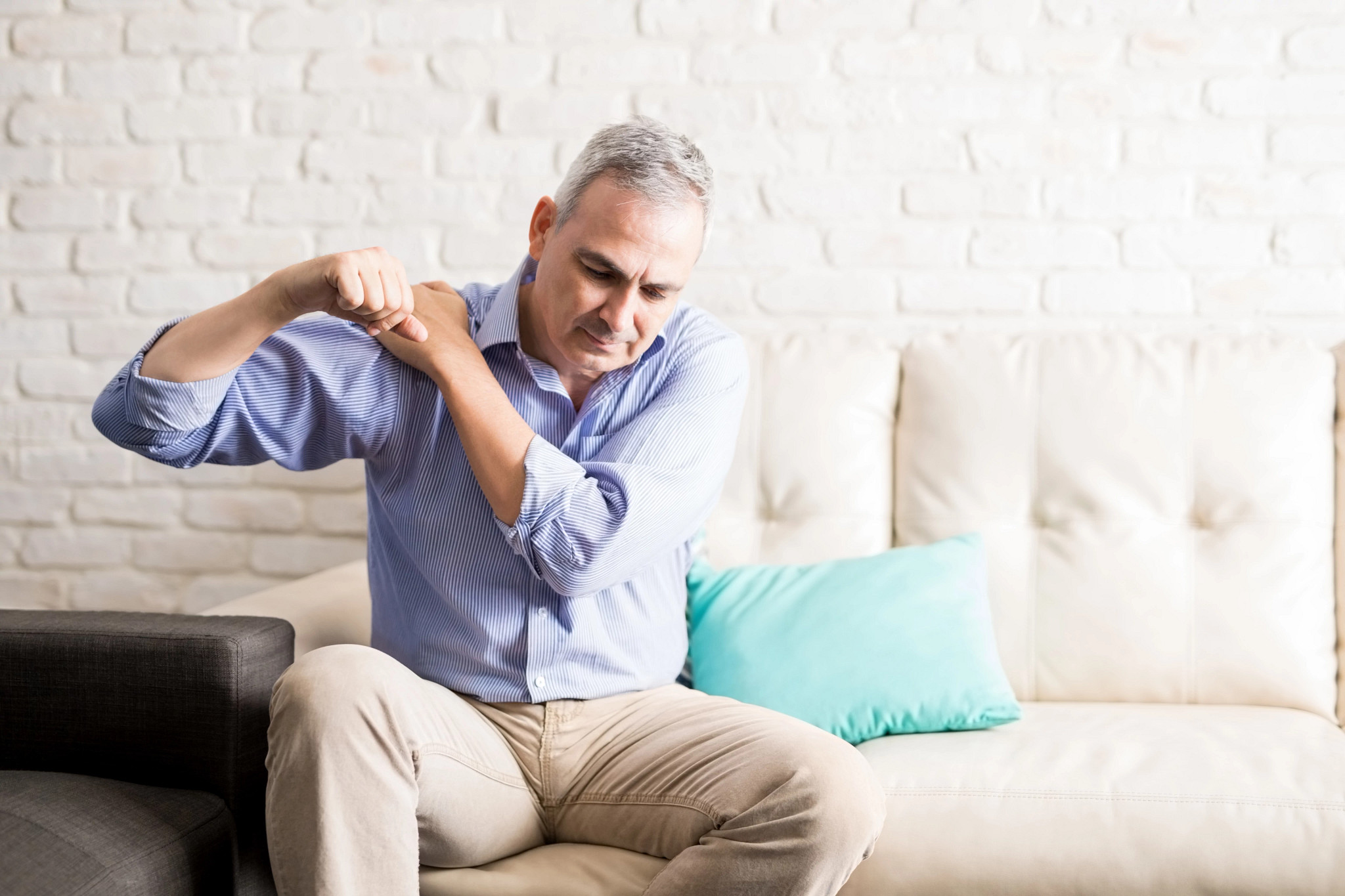 man sitting on the sofa and holding painful shoulder with another hand.