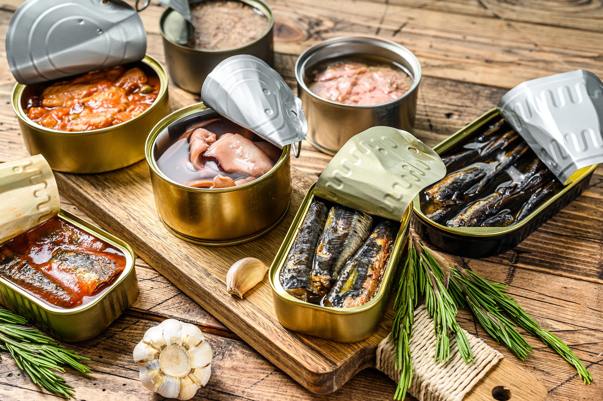 Open tins of sardines, tuna and other canned fish in oil and tomato sauce arranged on a rustic wooden table.