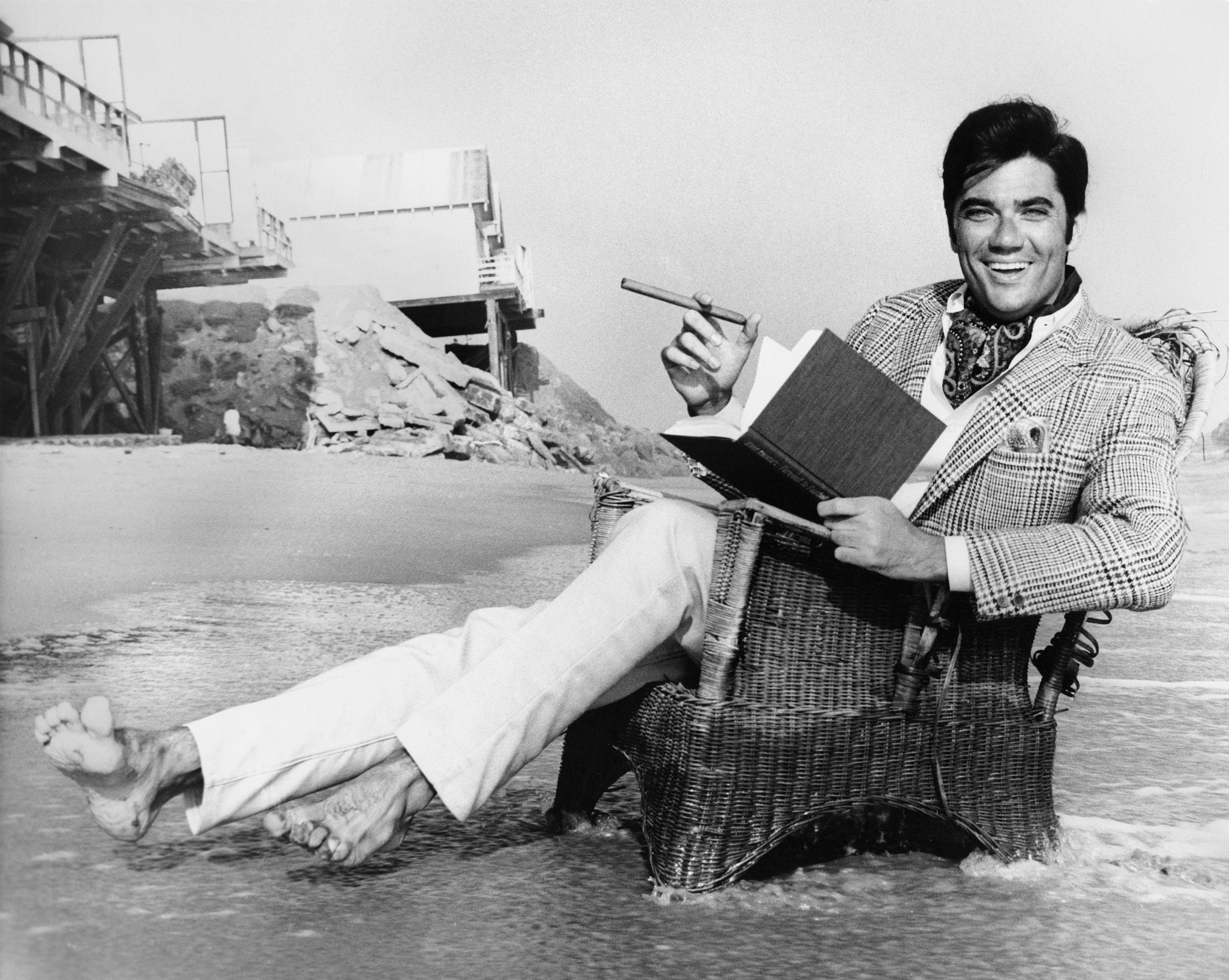 black and white image of rex reed reclining in a chair on a beach with a cigar and a book