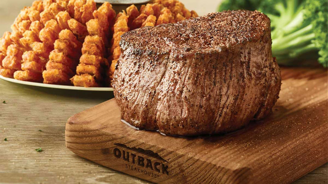 Outback Steakhouse steak filet on wood board with broccoli and bloomin onion in background