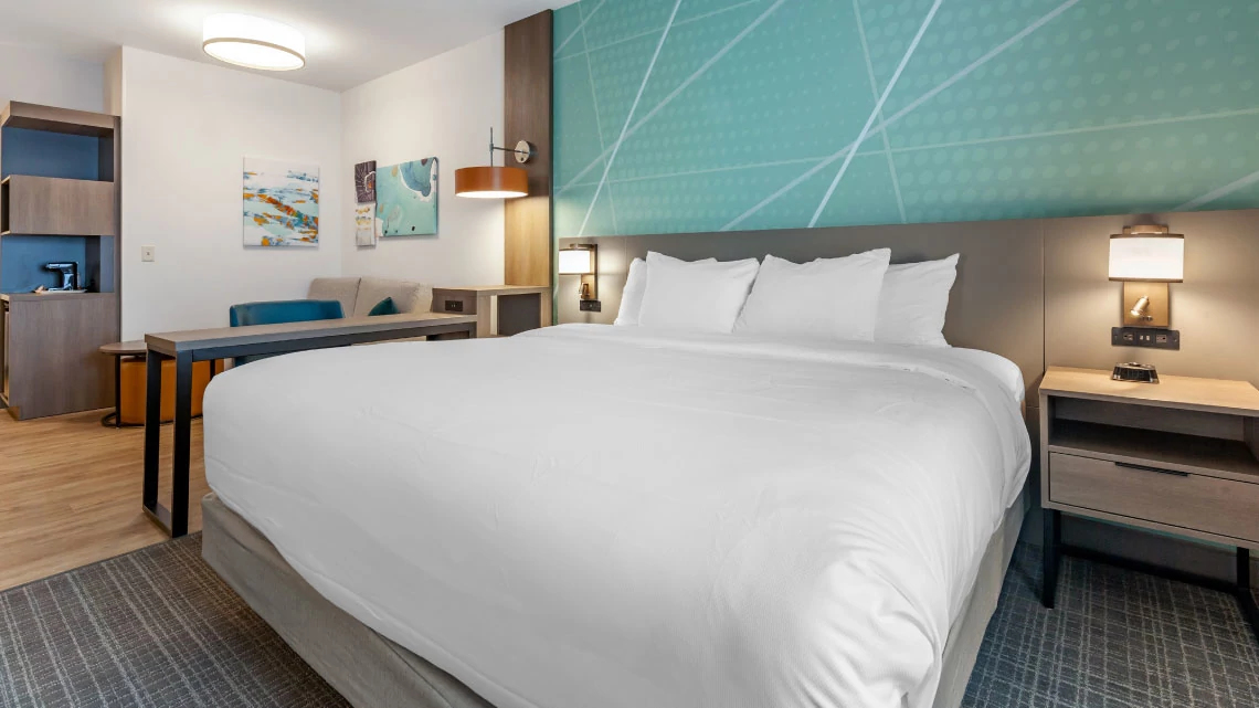 large bed with white linens with blue background, desk, coffee machine, multiple lamps in hotel room