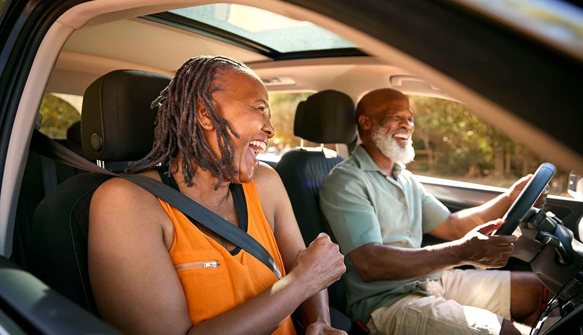 couple inside car smiling at sunset, man wearing short sleeves and shorts is driving and woman wearing an orange sleeveless blouse is in passenger seat, sunroof exposed