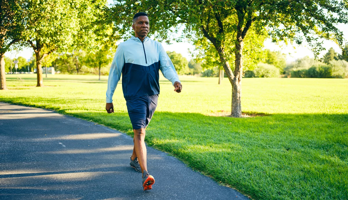 African American man with short hair wearing a light and dark blue athletic jacket and navy shorts walks along a paved path in a sunlit park