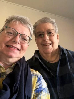 Close-up indoor selfie of two adults wearing glasses and layered casual clothing, standing side by side and looking toward the camera