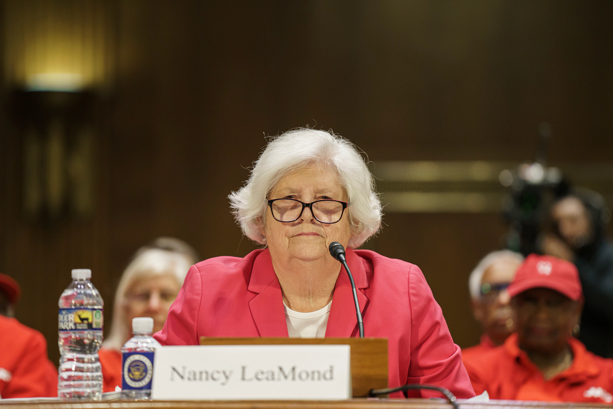 A woman in glasses, a red blazer and white shirt, sits in front of a microphone.