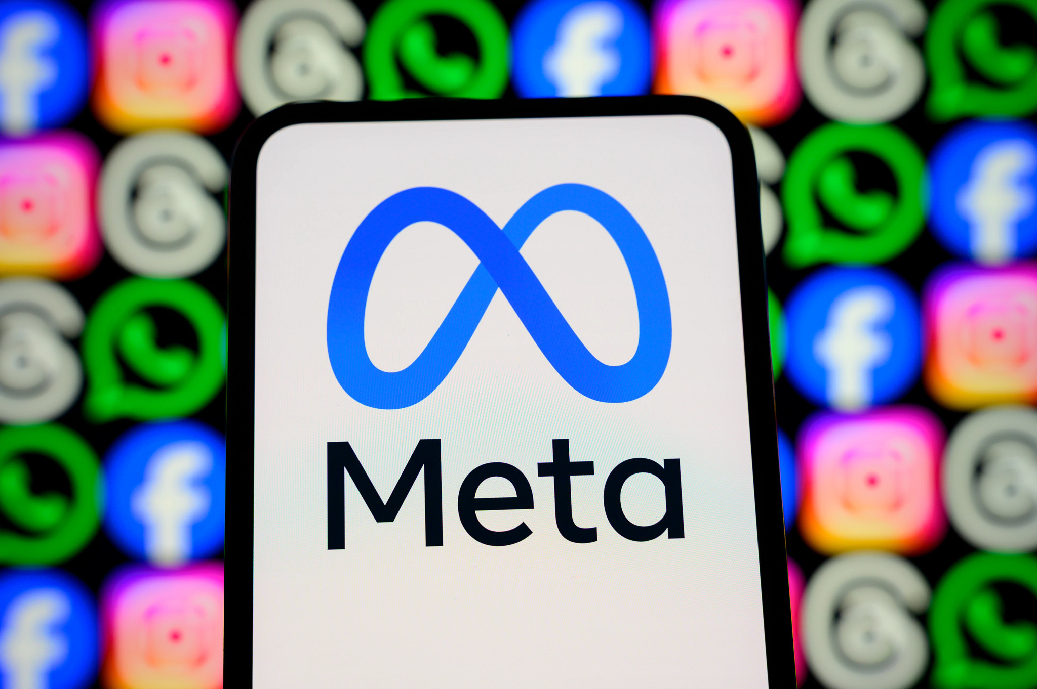 The meta logo appears on a smartphone screen in front of a screen showing icons for facebook, instagram, whats app and other meta apps