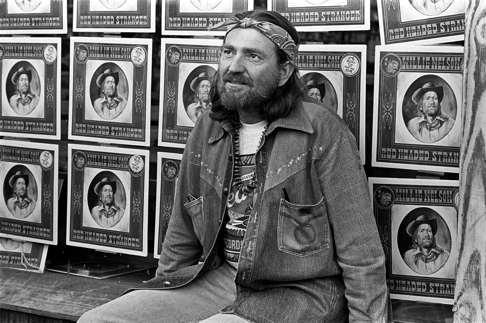 willie nelson sits on a bench in front of dozens of his red headed stranger album covers