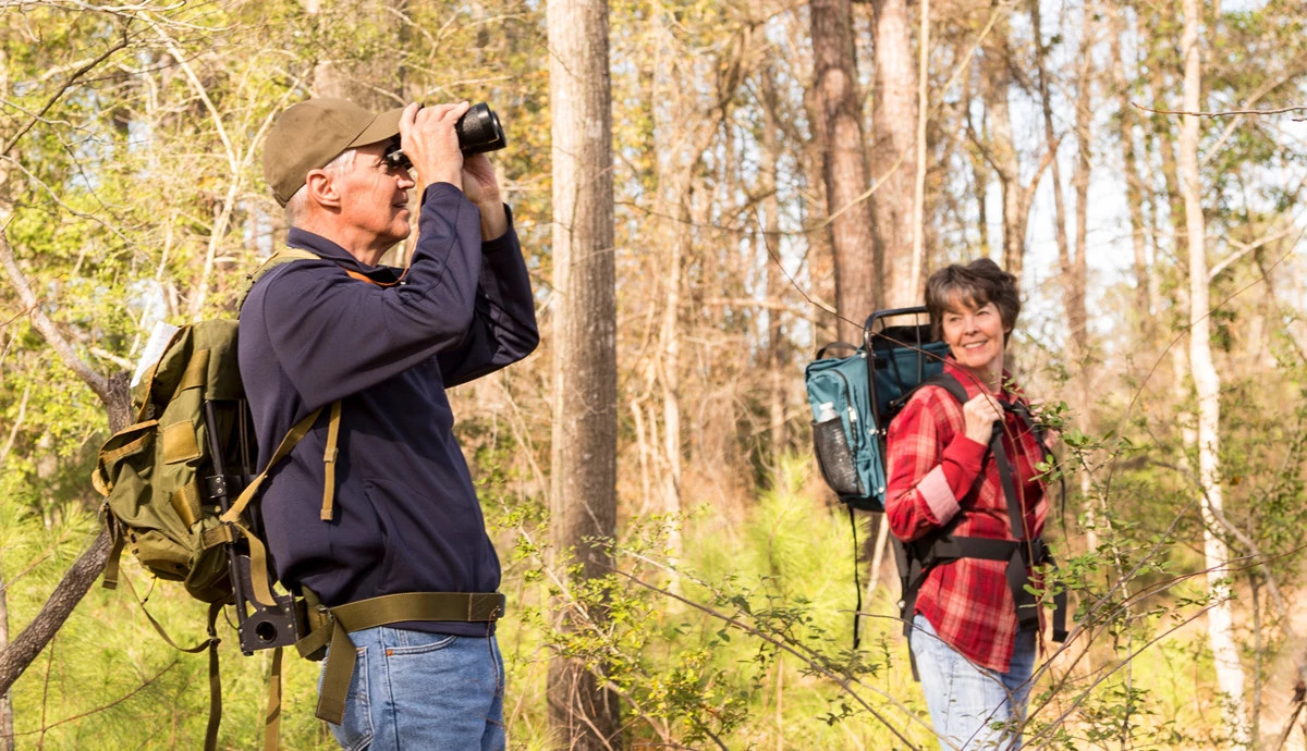 A man looks through binoculars while hiking outside with a companion.
