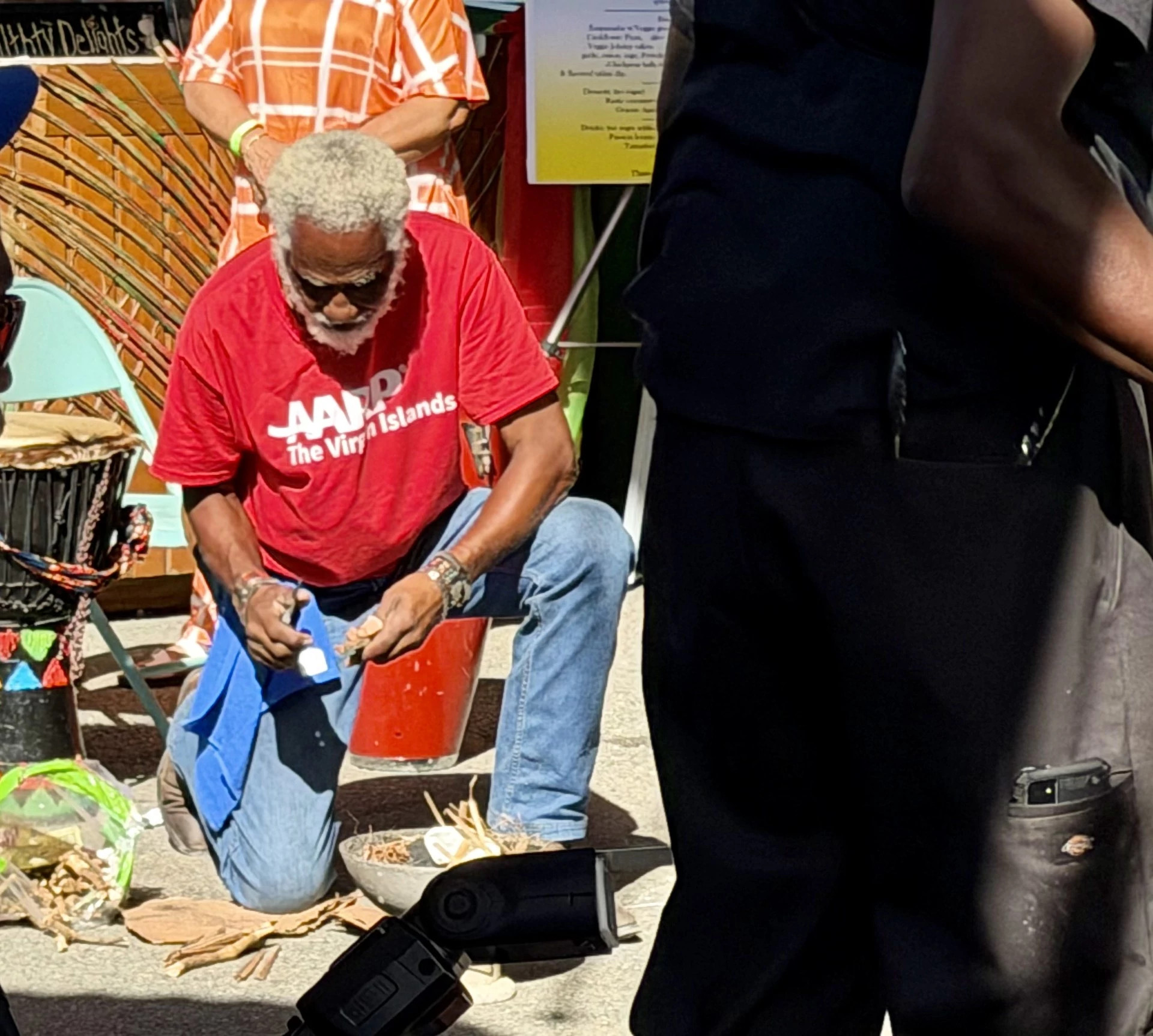 A man with gray hair, wearing a red "AARP Virgin Islands" t-shirt and sunglasses, kneels on the ground to light a traditional coal pot stove during a competition