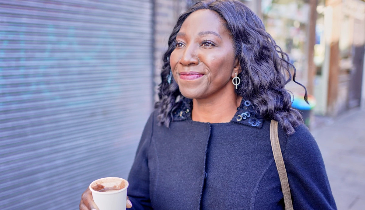 Mature woman smiling while walking down the street and holding a small coffee cup filled with coffee.