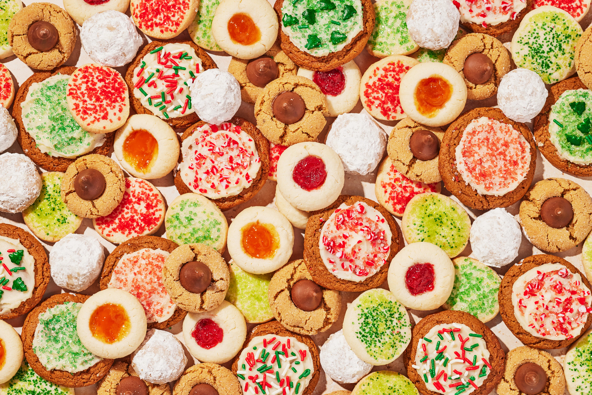 Overhead view of a colorful assortment of holiday cookies arranged closely together, including jam thumbprint cookies, powdered sugar snowball cookies, peanut butter blossom cookies, frosted cookies with festive red and green sprinkles and sugar cookies decorated for seasonal celebrations