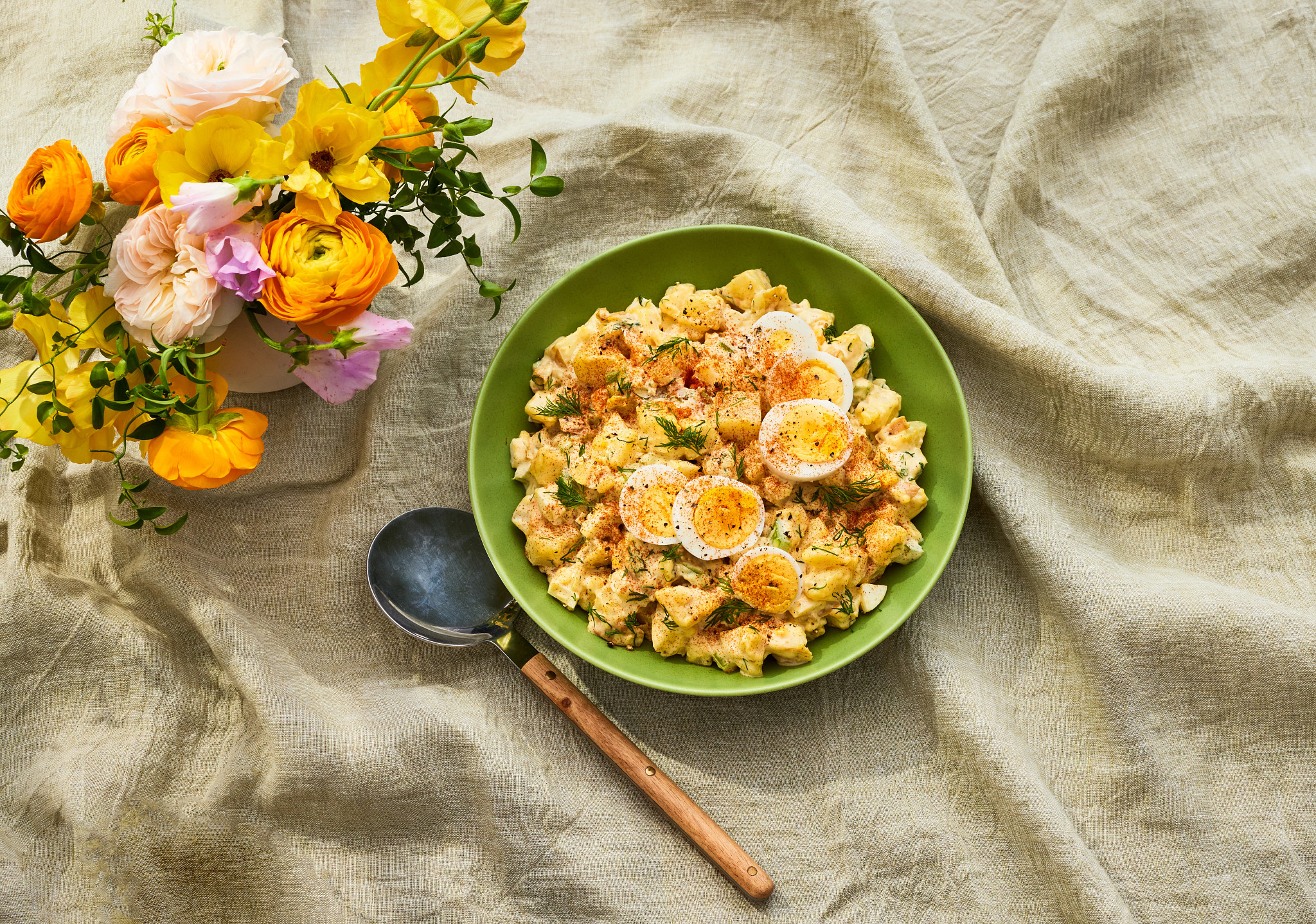 A bright green bowl filled with creamy egg salad topped with sliced hard-boiled eggs, fresh dill, and paprika, styled on a neutral linen cloth with a spoon and a vase of colorful spring flowers nearby