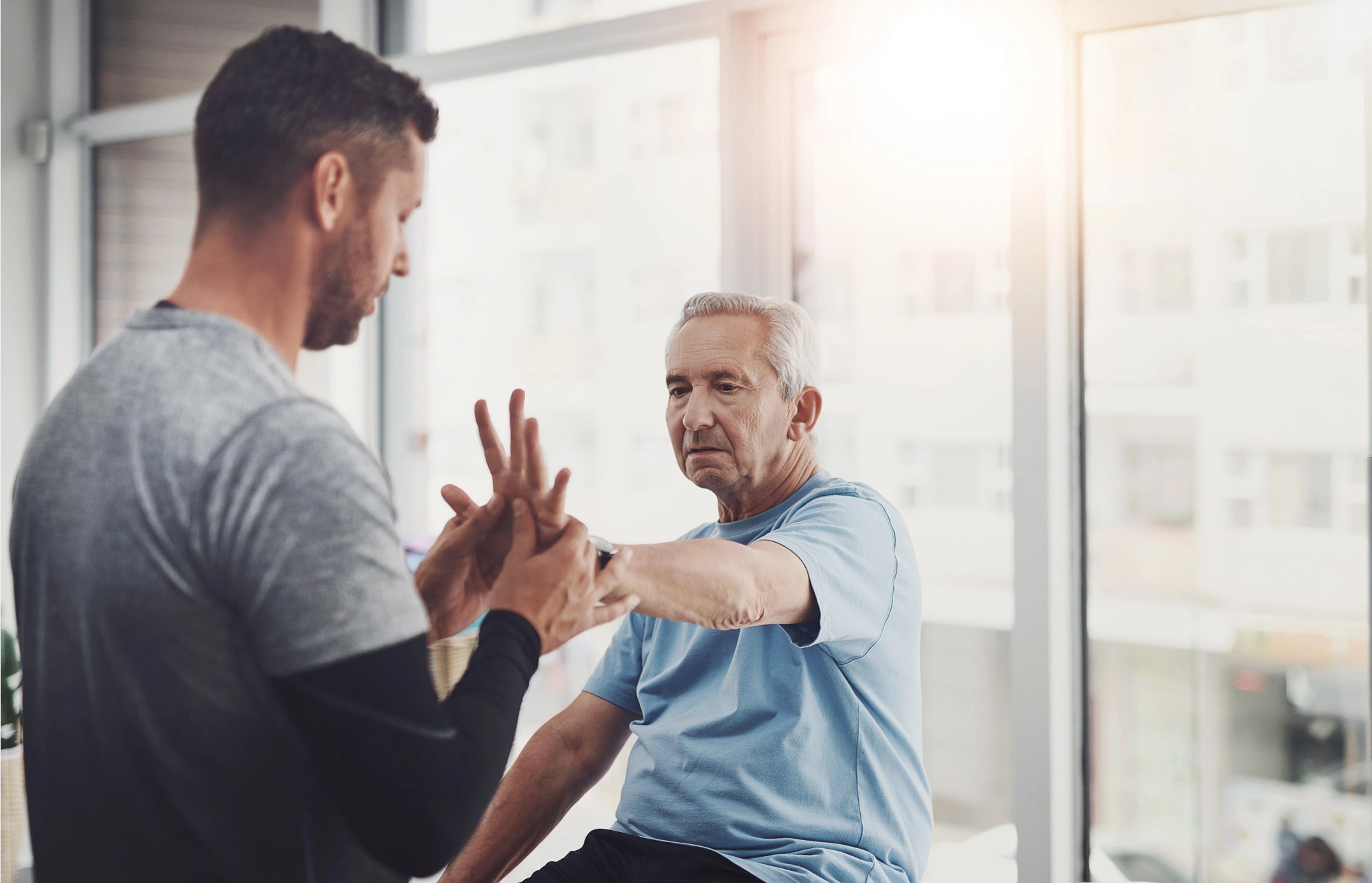 personal trainer or physical therapist in a gray shirt guides an older man in a blue t-shirt through a hand and arm mobility exercise