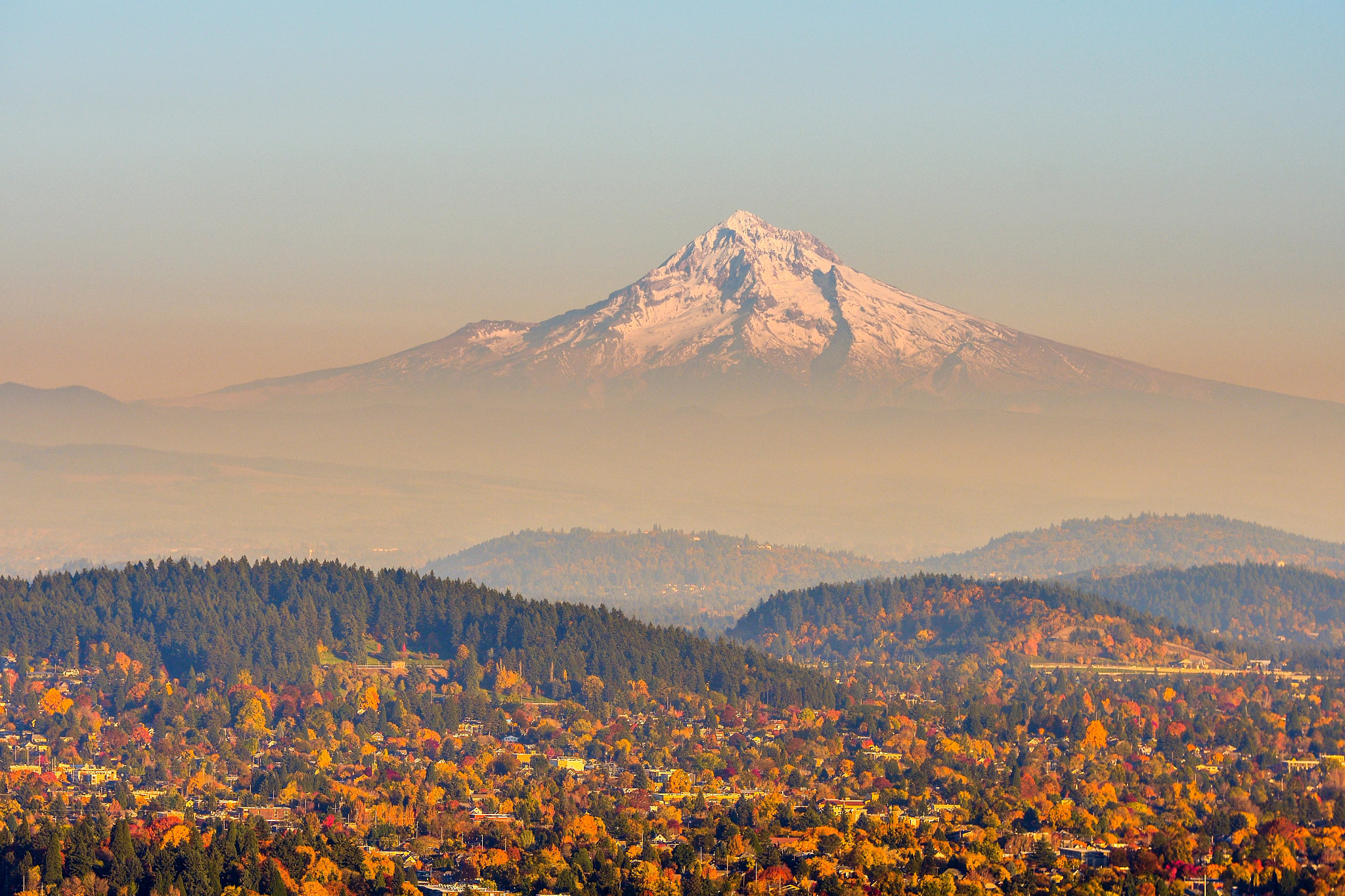 Snow-capped mountain towering above a colorful autumn landscape with forests, rolling hills, and a small community below