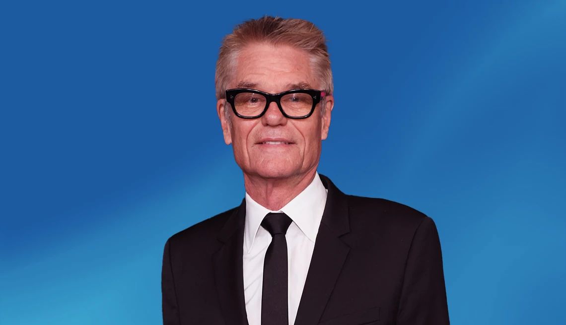 Harry Hamlin in a black suit and tie against a blue ombre background