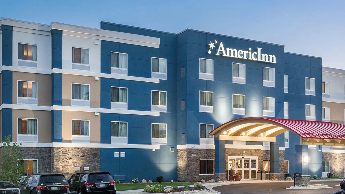 AmericInn® by Wyndham Front view of the AmericInn hotel property, blue and white exterior, early evening, lights on, cars in parking lot