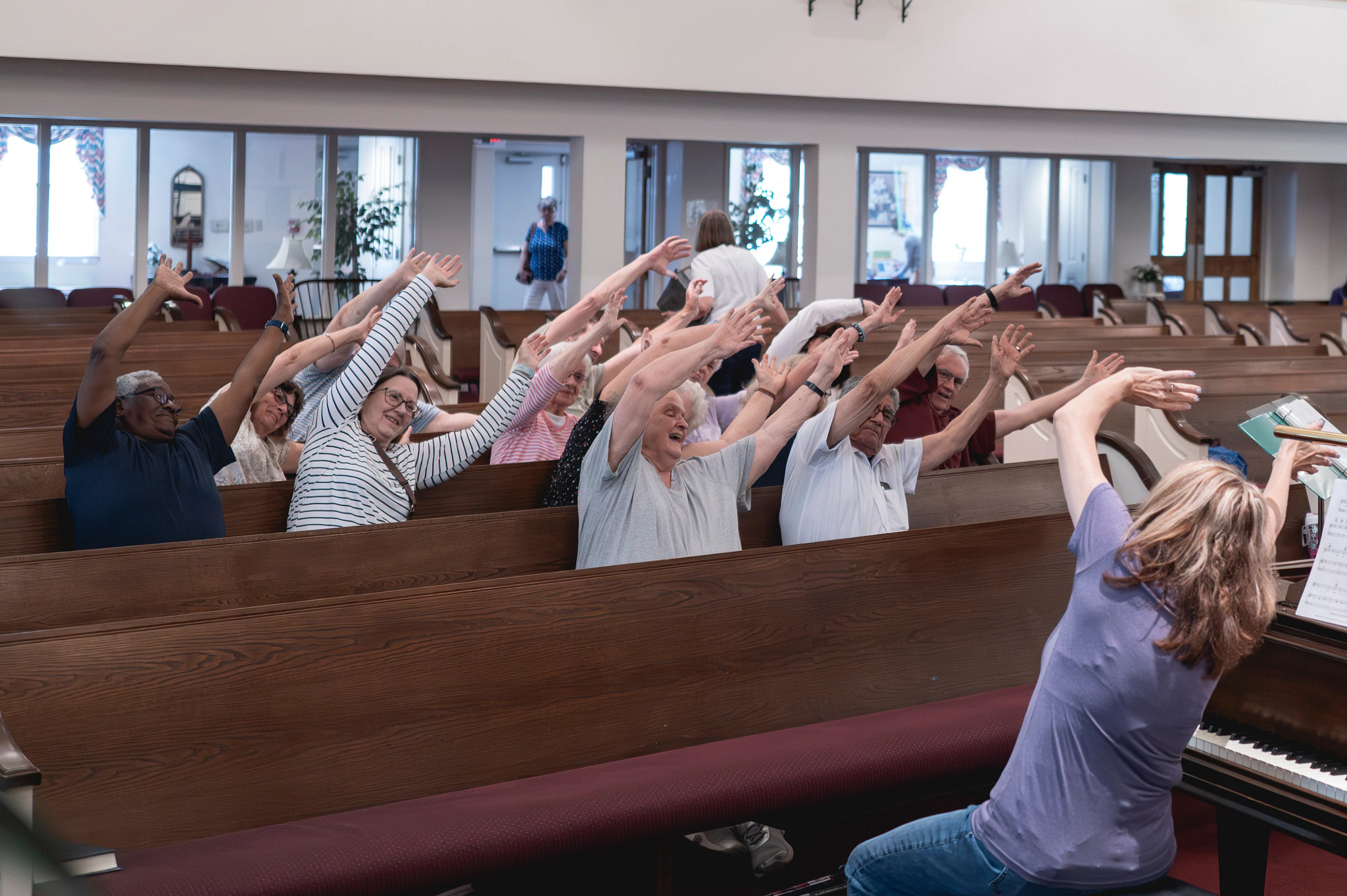 A photo shows Susan Dommer leading choir members in breathing and physical exercises at the start of rehearsal at Church of the Brethren in Manassas, Virginia.