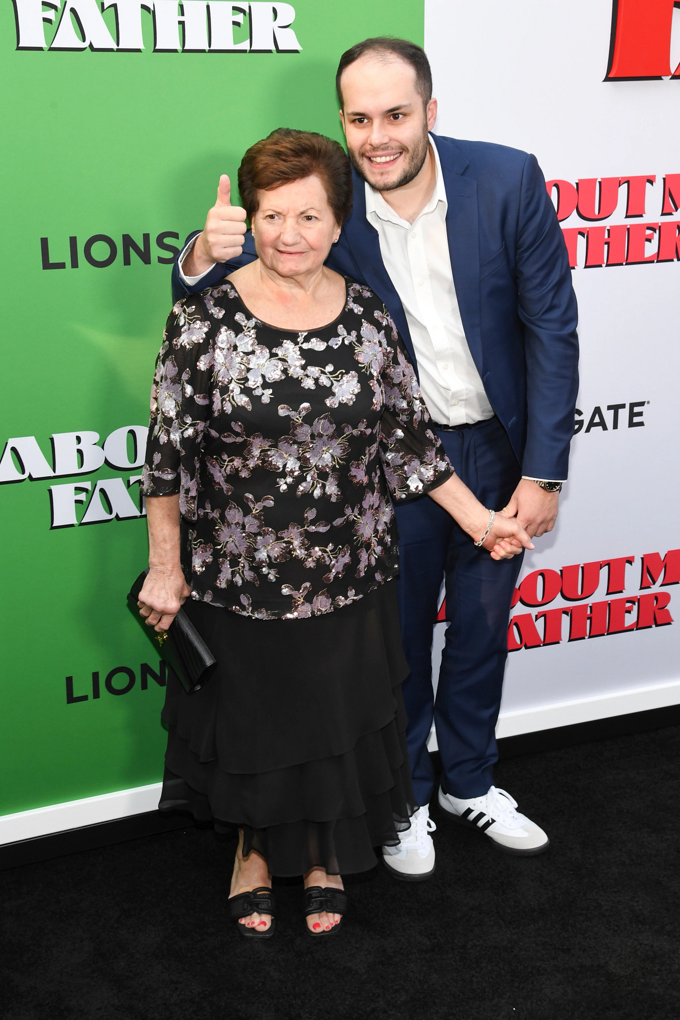 Nonna Gracie with her grandson Matt Gresia pose together on an event arrival carpet in front of a promotional backdrop. Matt gives a thumbs-up gesture while standing beside Nonna Gracie