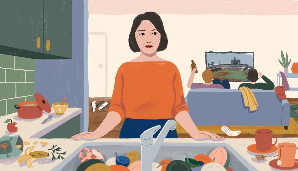 My Grown Sons Moved Back Home a woman stands at a sink full of dirty dishes while two men sit on the couch watching television behind her