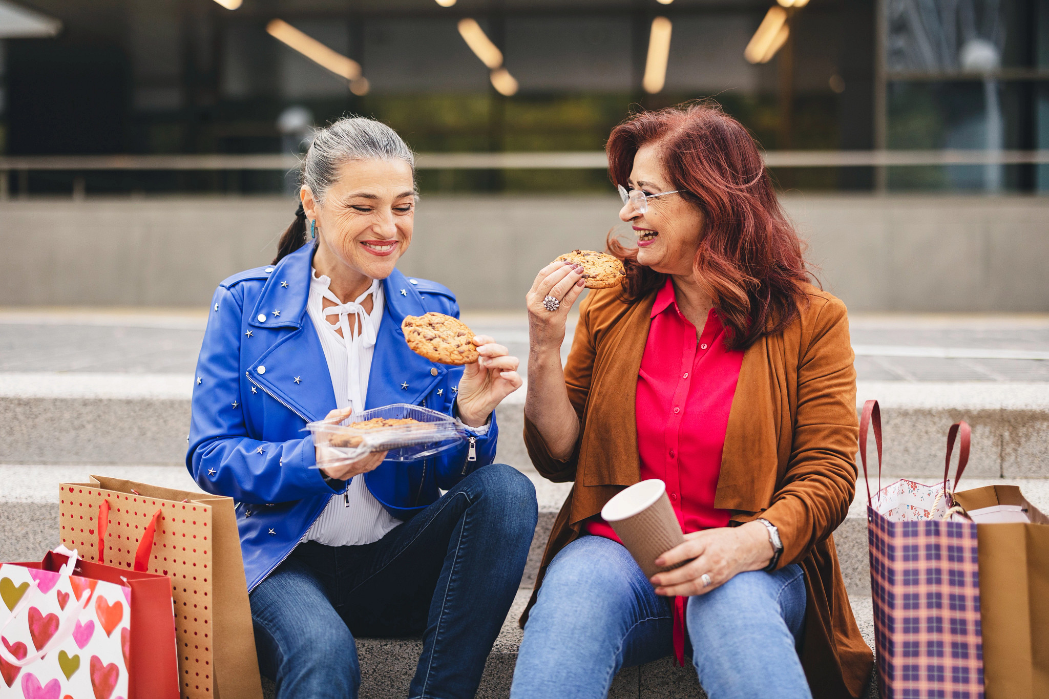 a photo shows two older adult women taking a break from shopping. They are outside, seated on concrete steps enjoying chocolate chip cookies