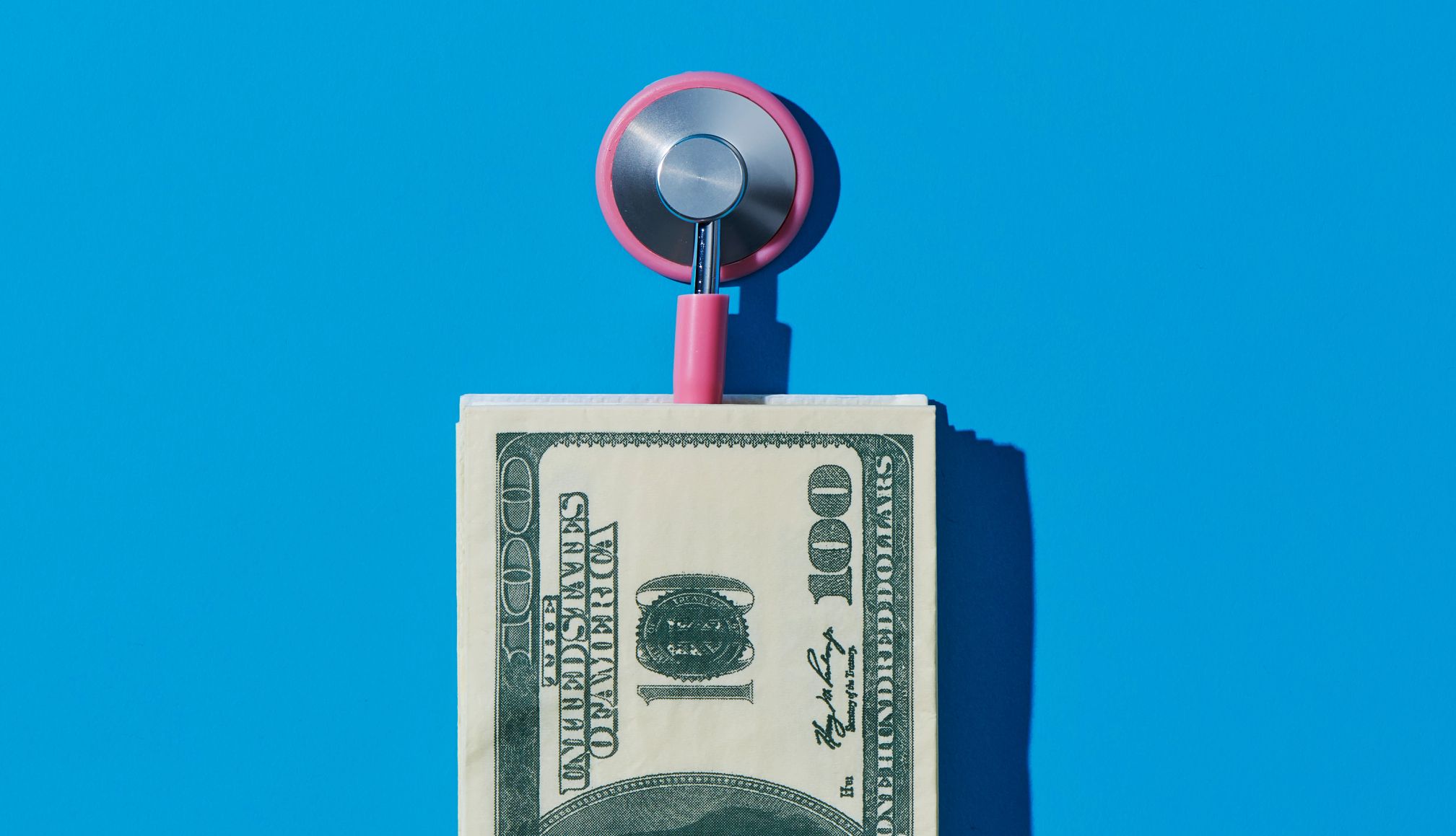 Un billete de 100 dólares adjunto a un estetoscopio, contra un fondo azul.